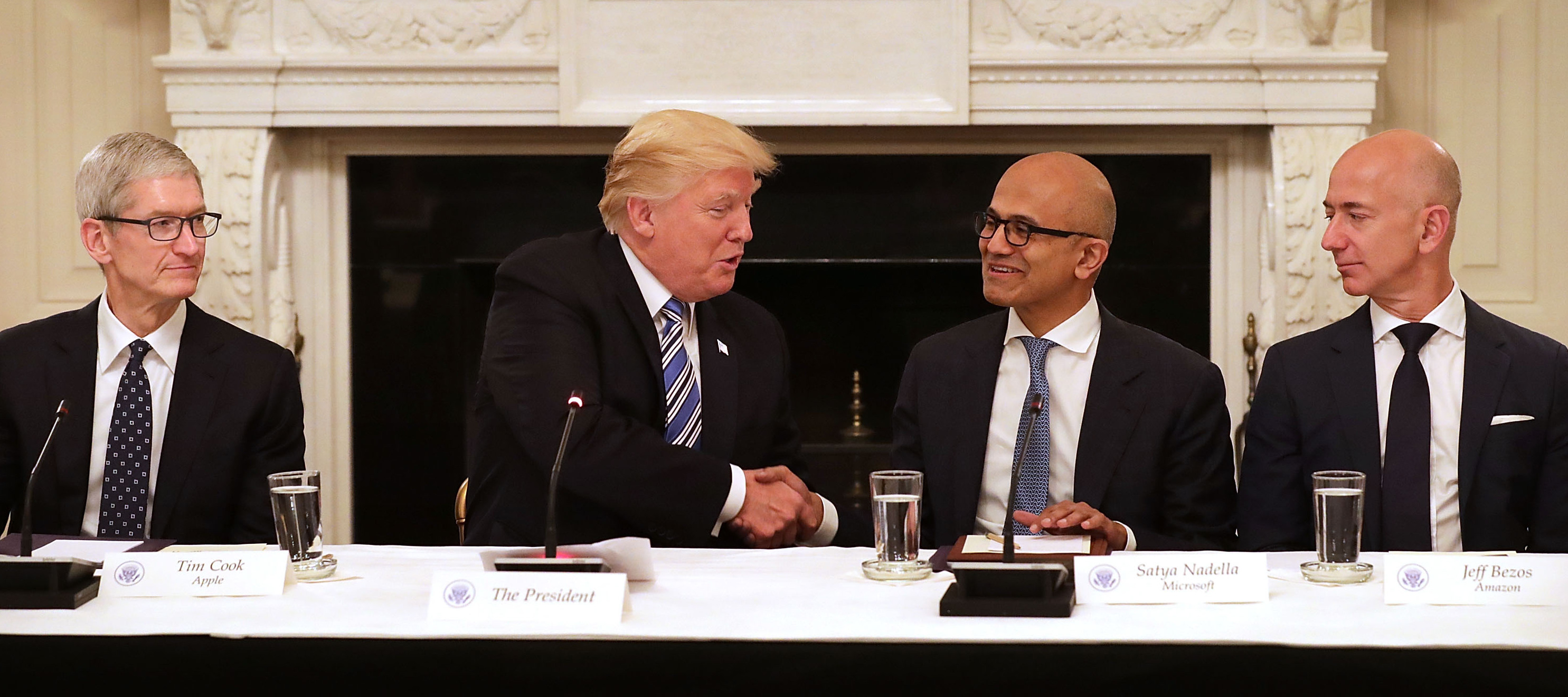 President Donald Trump shakes hands with Microsoft CEO Satya Nadella while sitting at a table with Apple CEO Tim Cook and Amaazon CEO Jeff Bezos.
