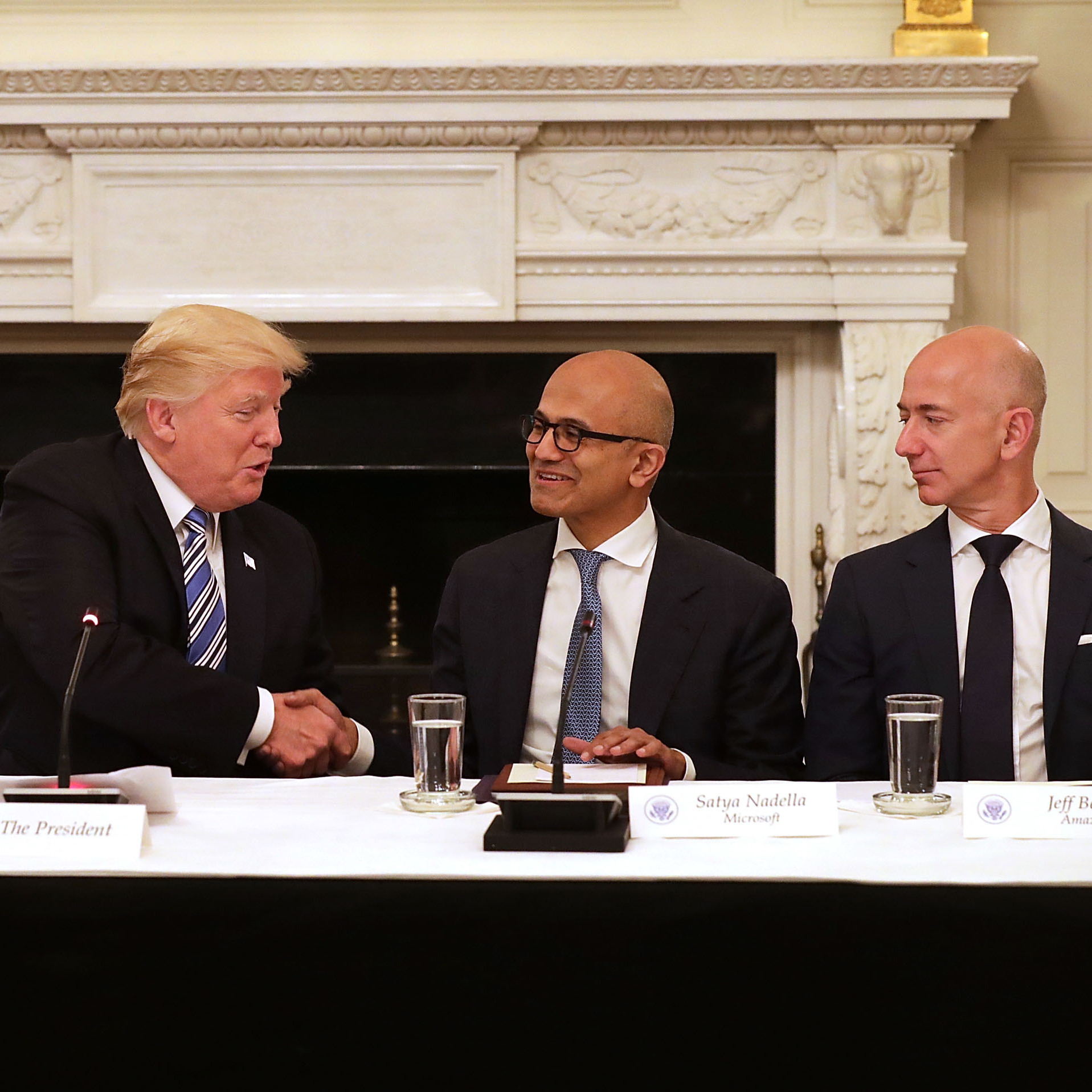 President Donald Trump shakes hands with Microsoft CEO Satya Nadella while sitting at a table with Apple CEO Tim Cook and Amaazon CEO Jeff Bezos.