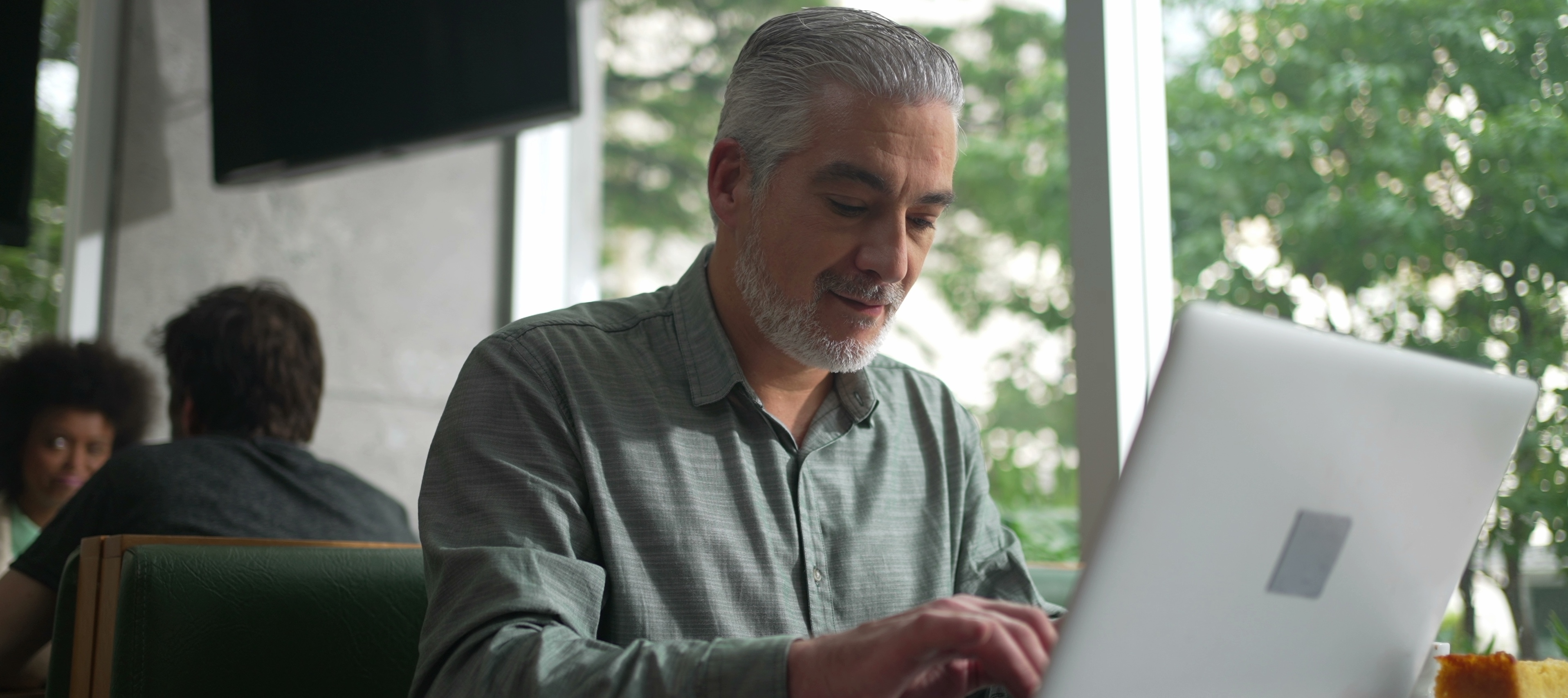 An attractive gray-haired bearded man works on his computer in a coffee shop.