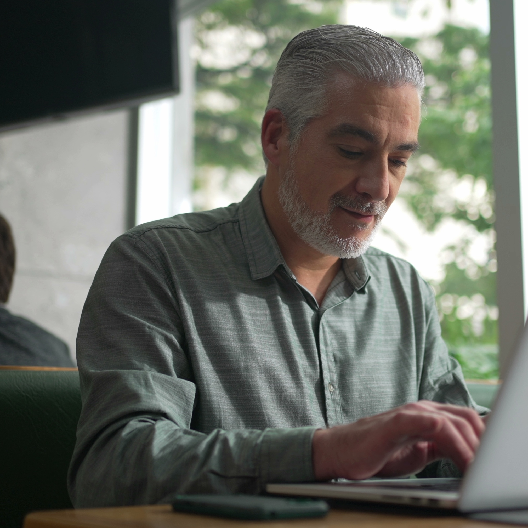 An attractive gray-haired bearded man works on his computer in a coffee shop.
