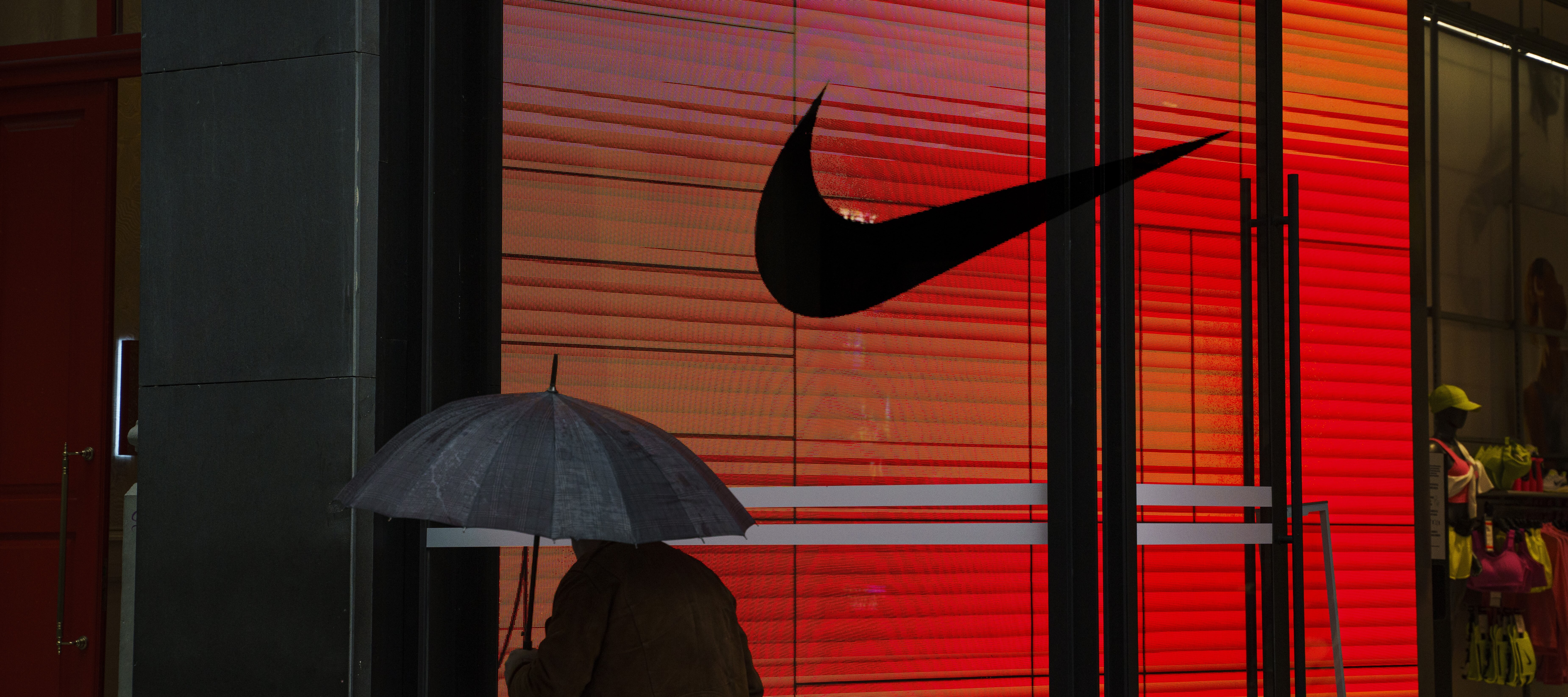 Nike, its logo scene here on a storefront as a person carrying an umbrella walks past, recently made its second round of job cuts in 2026, laying off 1,400 employees..