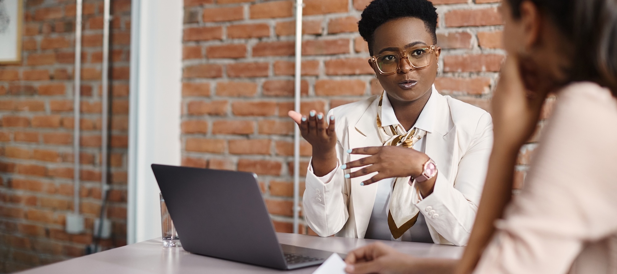A financial advisor speaks to her client, sitting at a desk with a laptop and paperwork