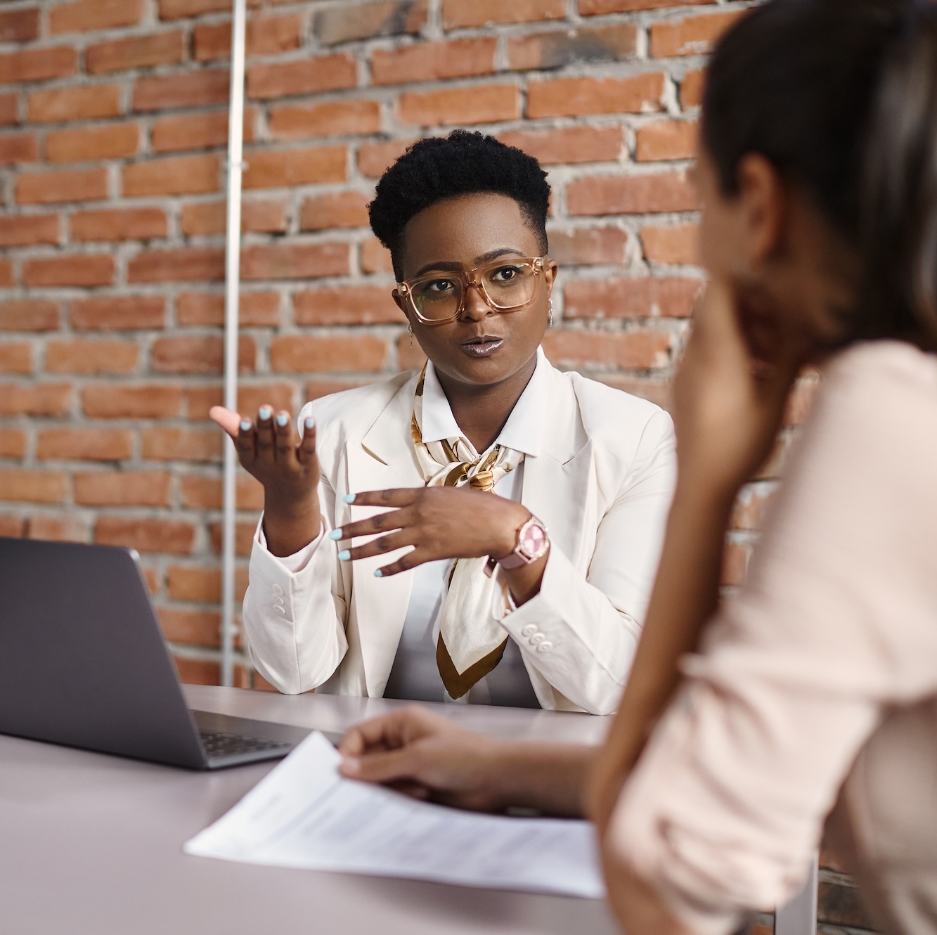 A financial advisor speaks to her client, sitting at a desk with a laptop and paperwork