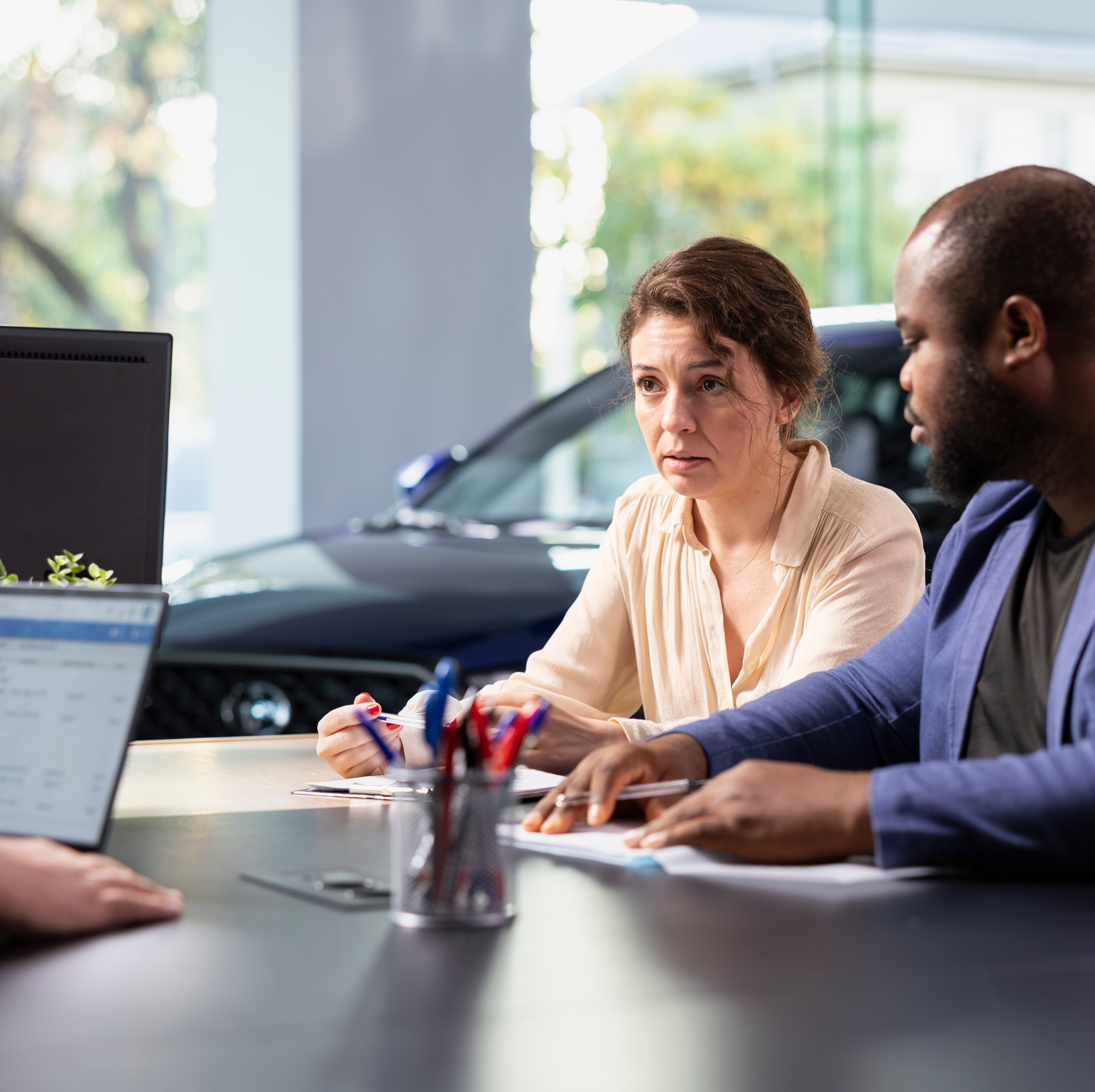 A couple in a car showroom talk to a car salesman about pricing on a car.