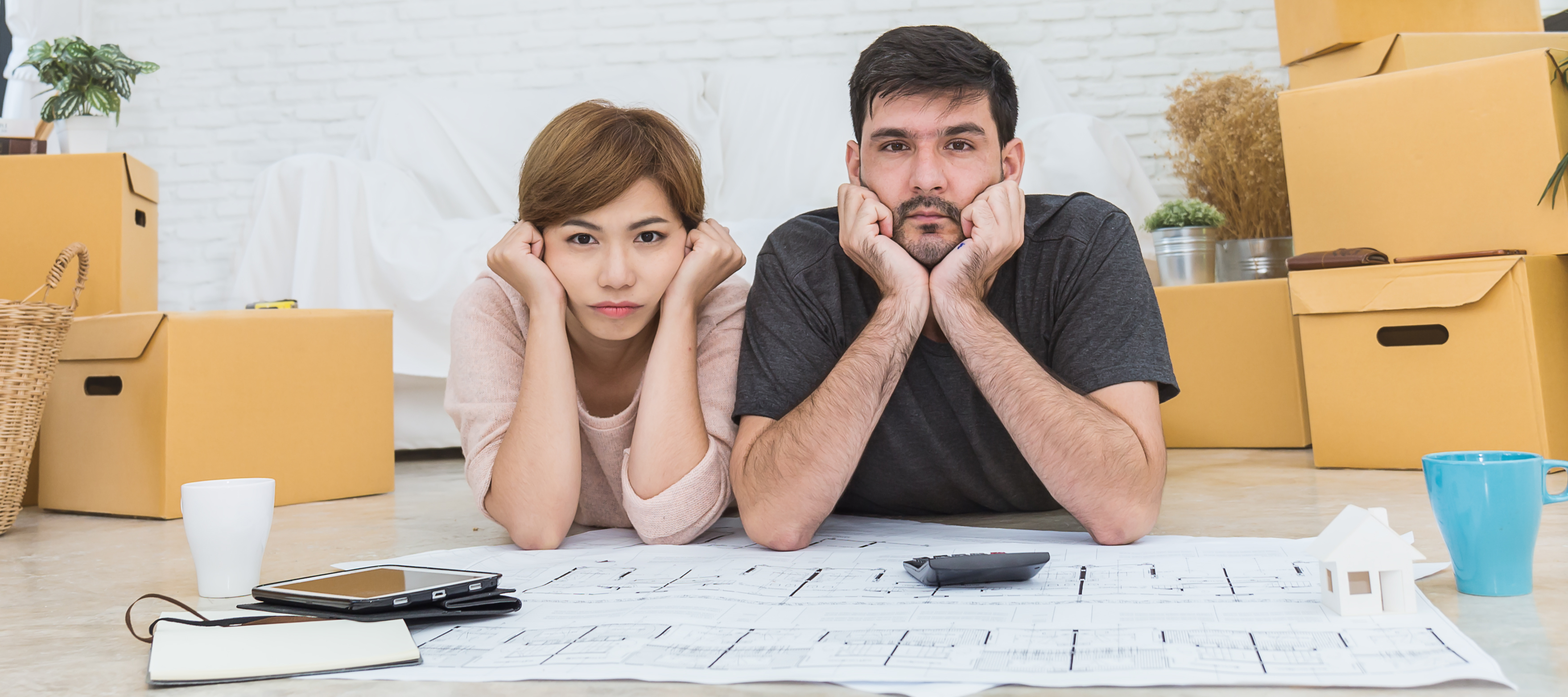A man and a woman lie on the floor looking sadly toward the camera amidst a room filled with cardboard boxes and house plans on paper.