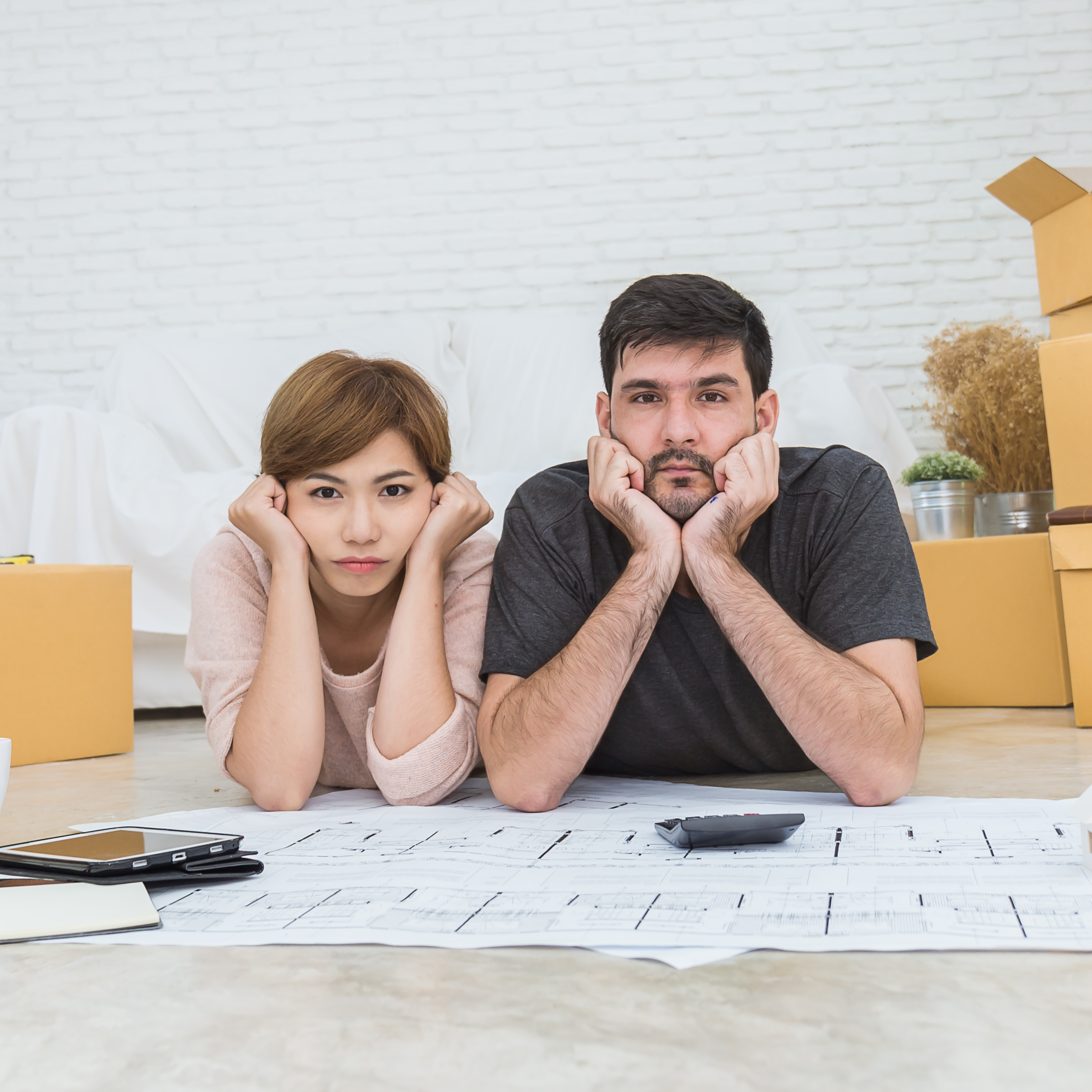A man and a woman lie on the floor looking sadly toward the camera amidst a room filled with cardboard boxes and house plans on paper.