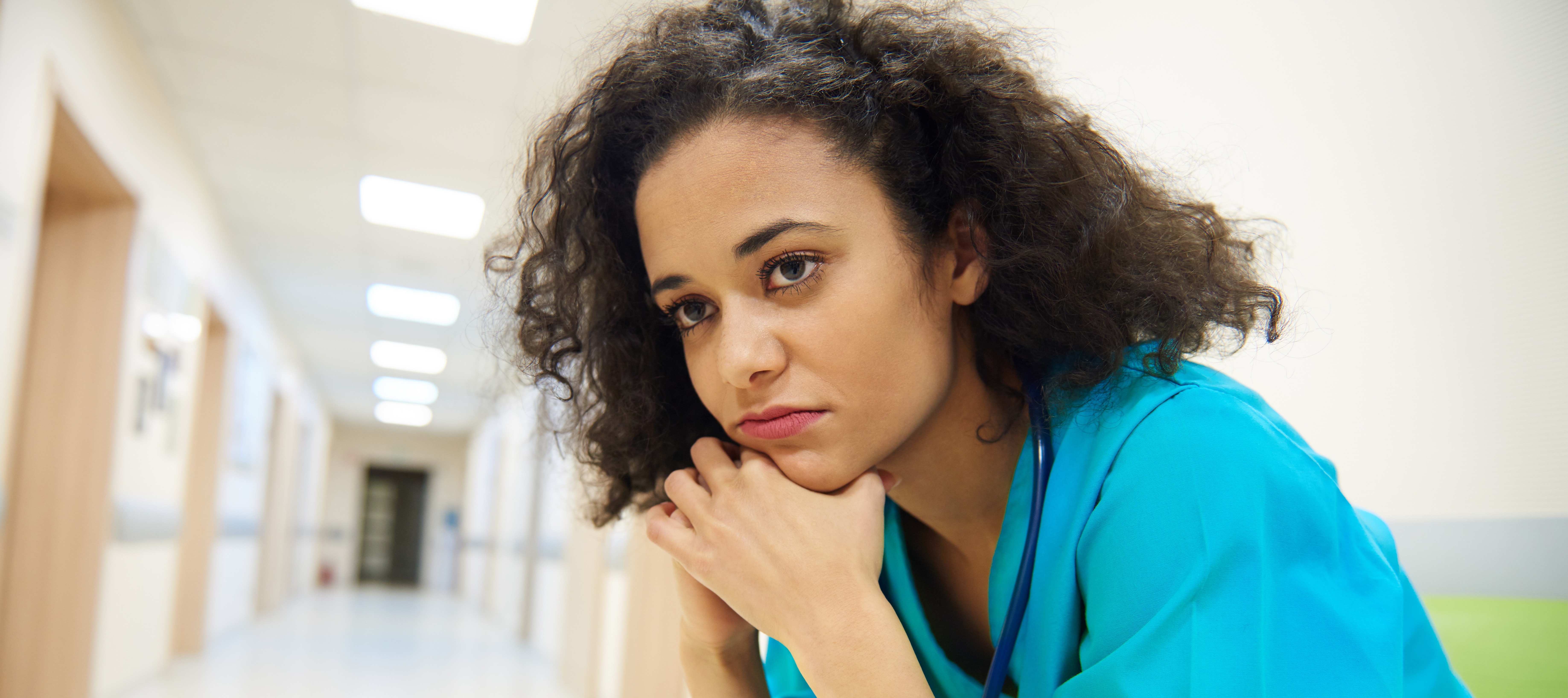 A woman wearing blue scrubs is sitting on a chair in a bright hallway, head resting in her hands, in a moment of introspection.