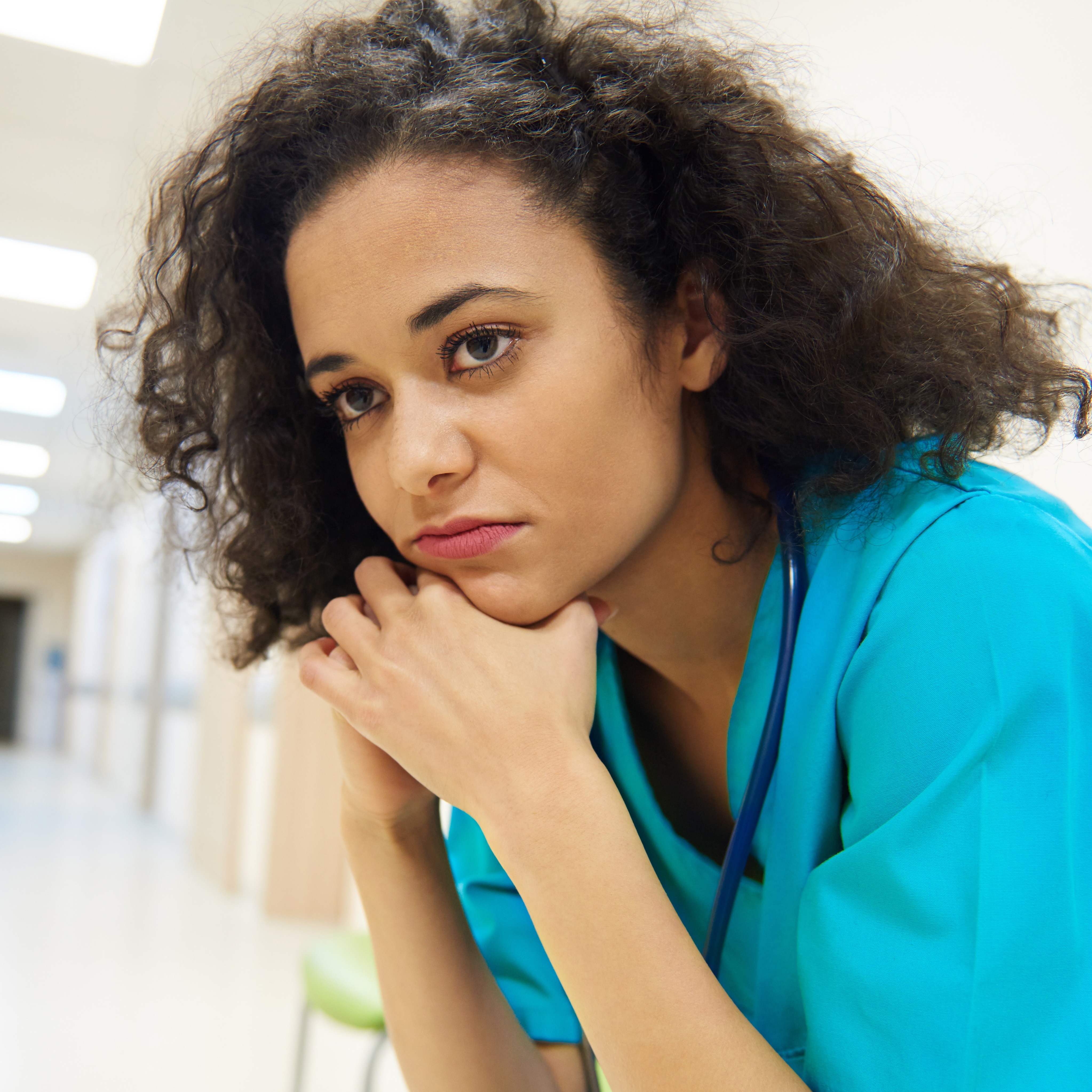 A woman wearing blue scrubs is sitting on a chair in a bright hallway, head resting in her hands, in a moment of introspection.