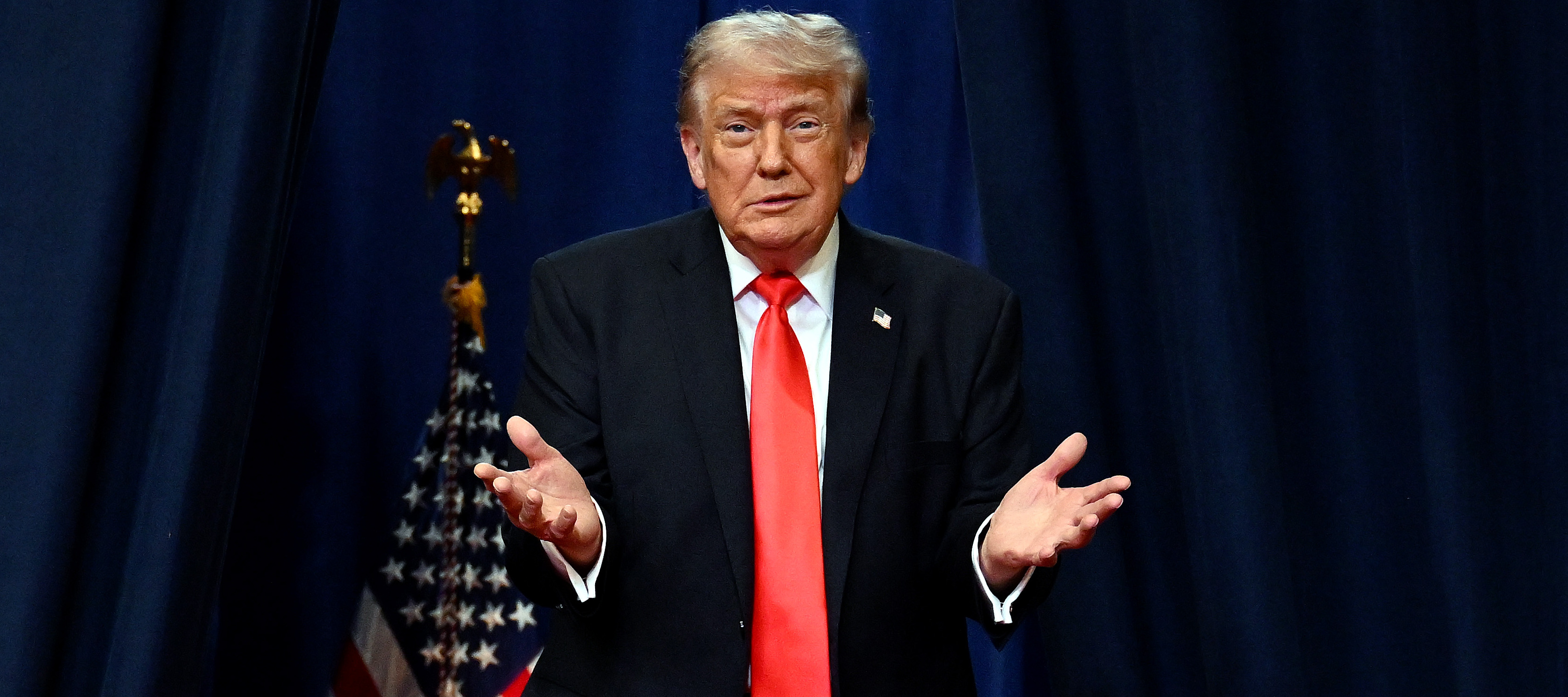 President Donald Trump stands on a stage with a dark curtain behind him and an American flag, holding his hands up and shrugging his shoulders.