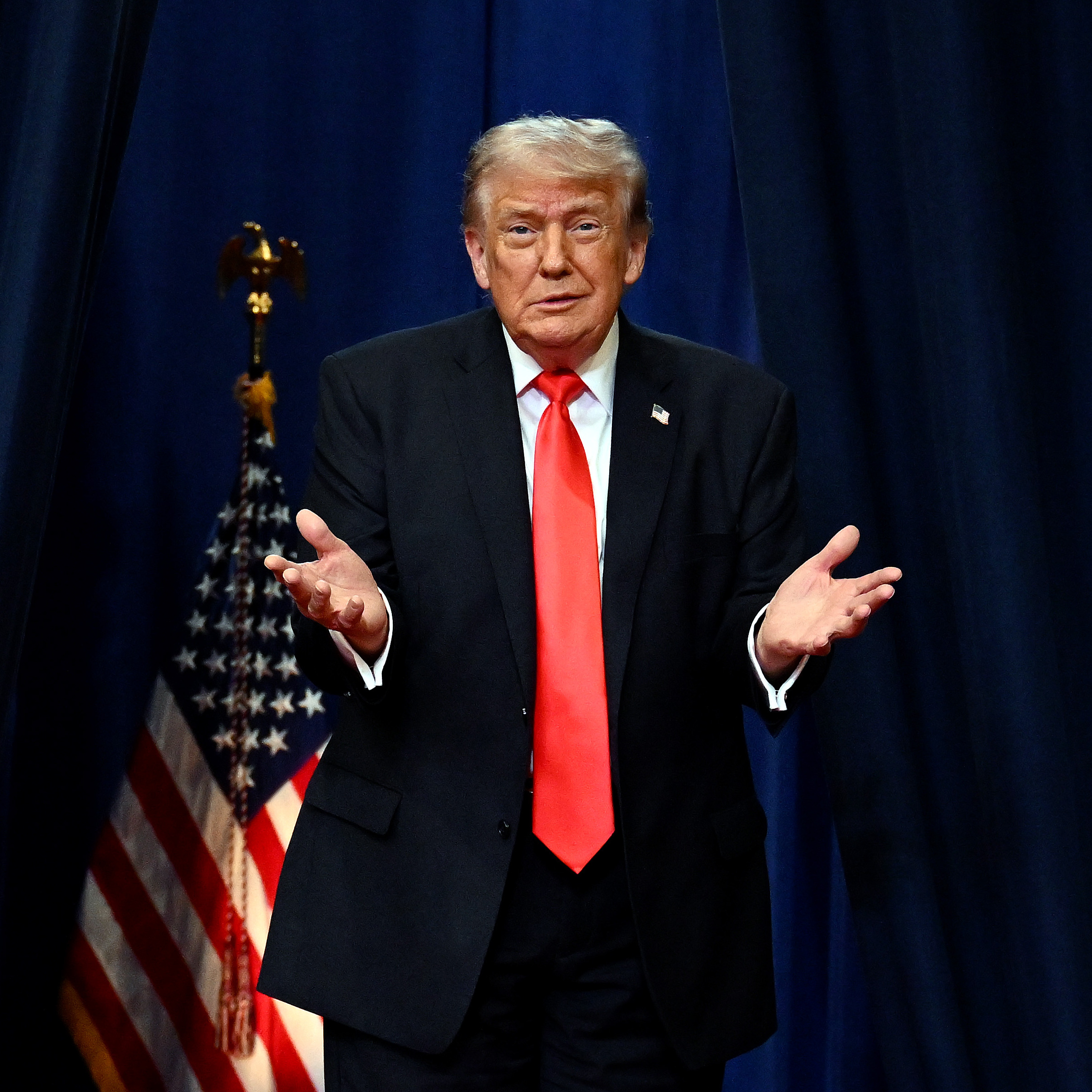 President Donald Trump stands on a stage with a dark curtain behind him and an American flag, holding his hands up and shrugging his shoulders.