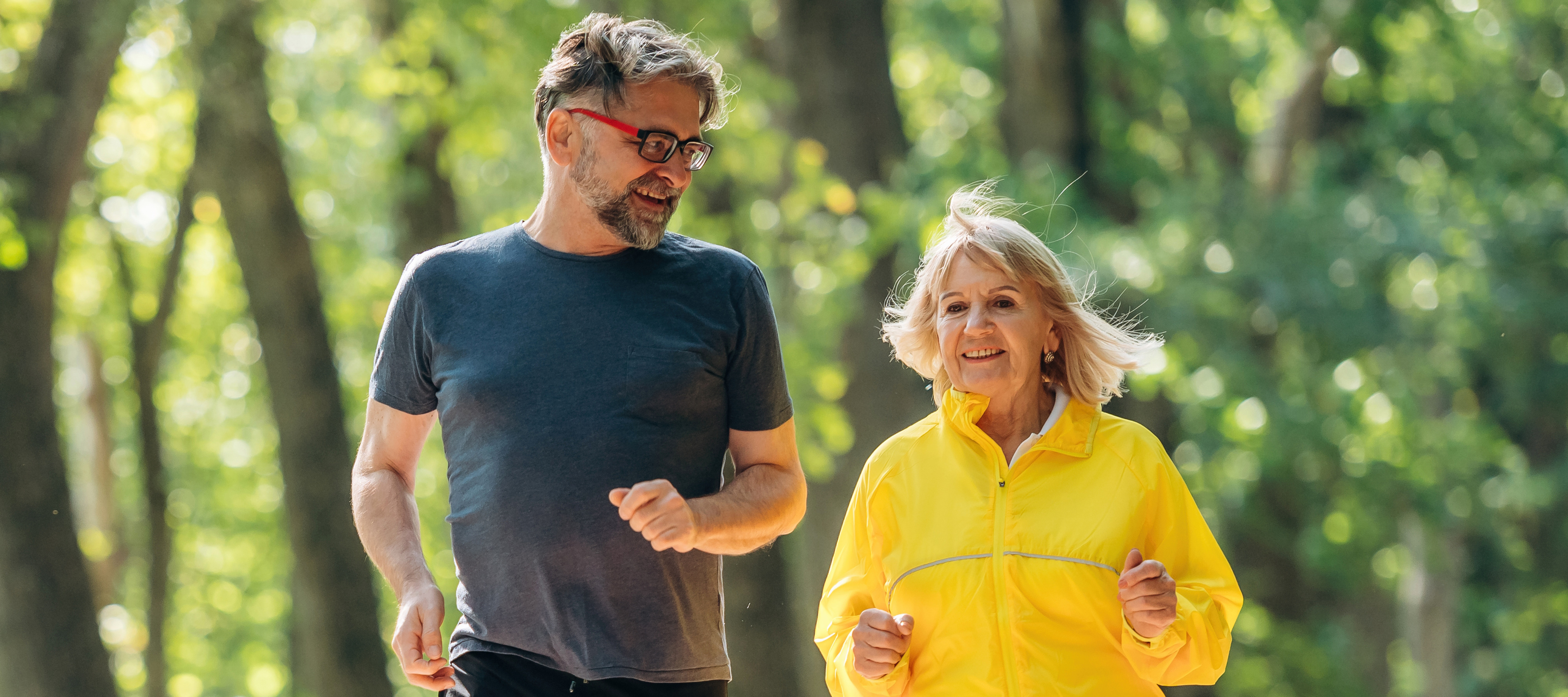 An older couple run down a wooded forest path together.