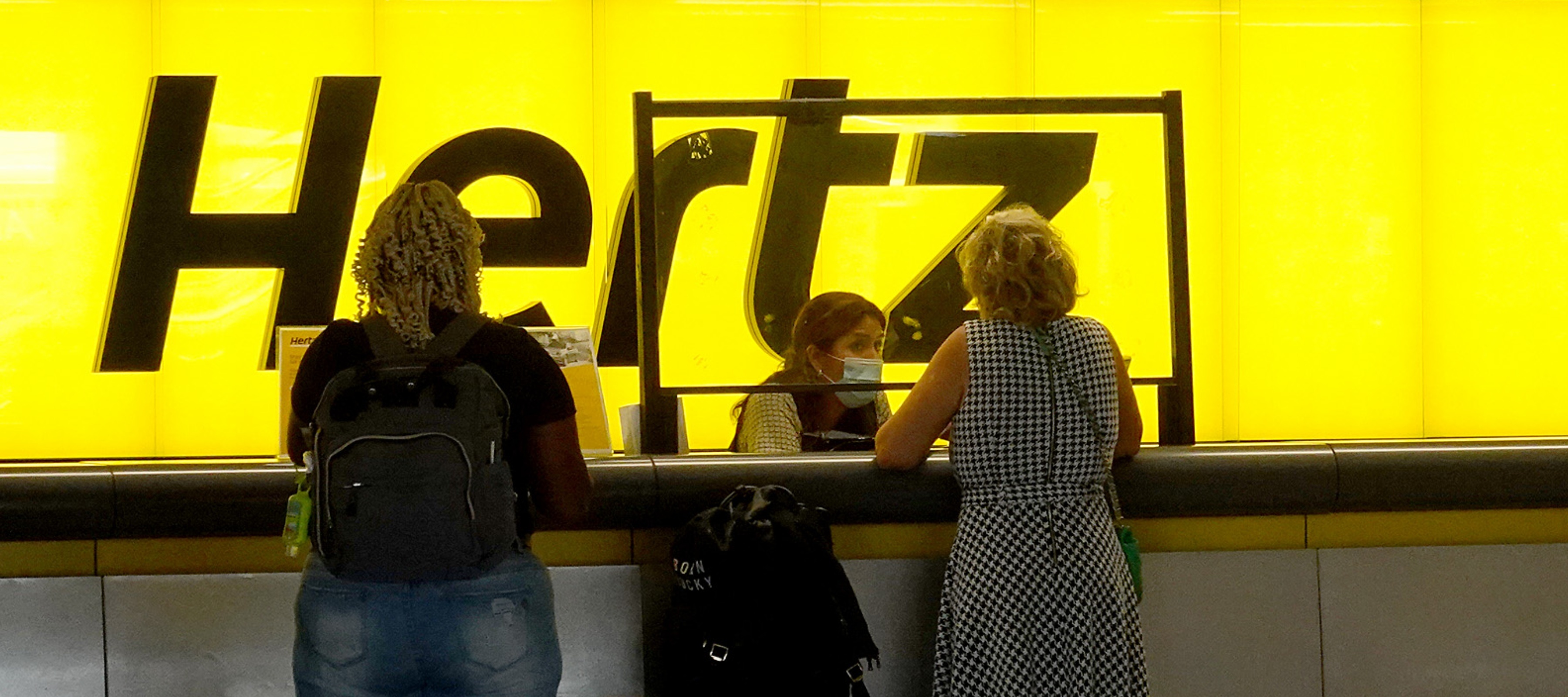 Three people stand in front of a brightly lit Hertz counter at an airport