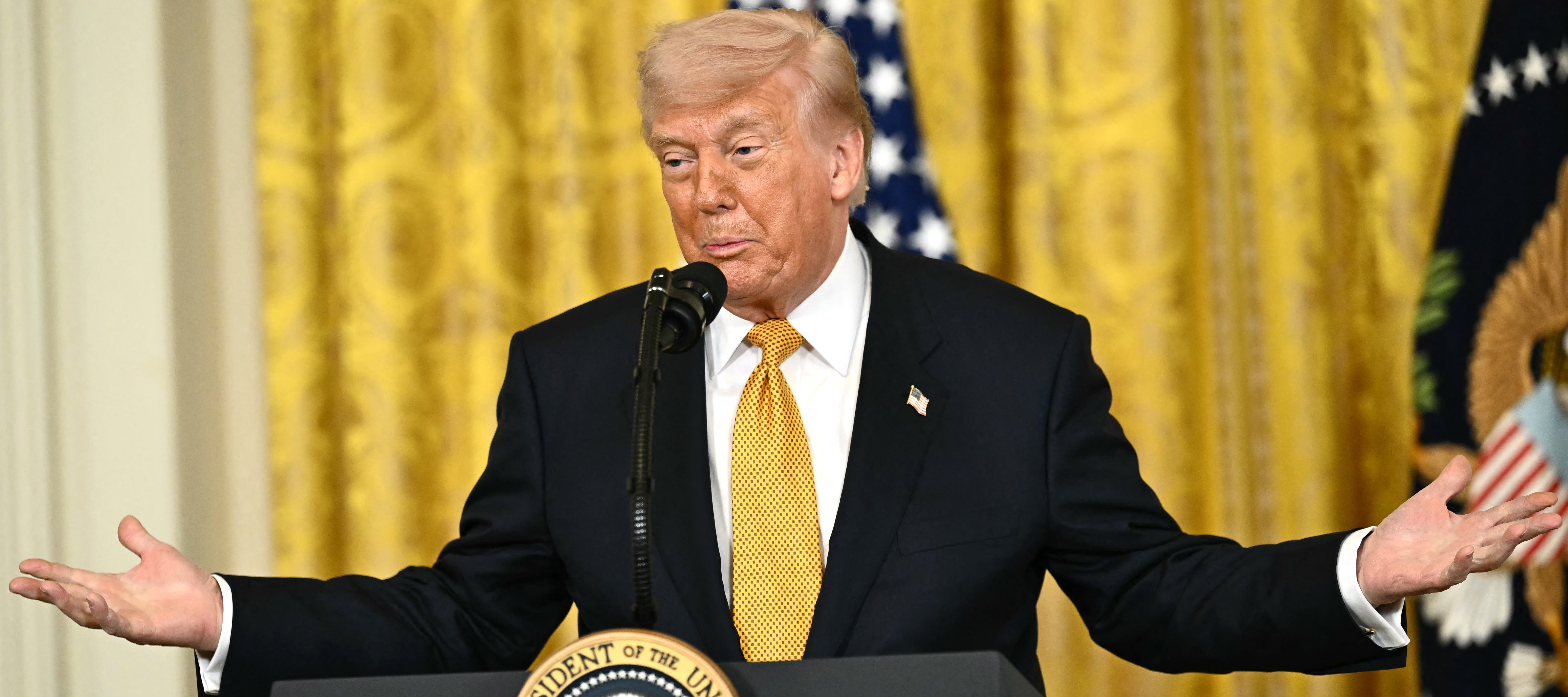 President Donald Trump spread his arms wide and shrugs against a golden backdrop on stage.