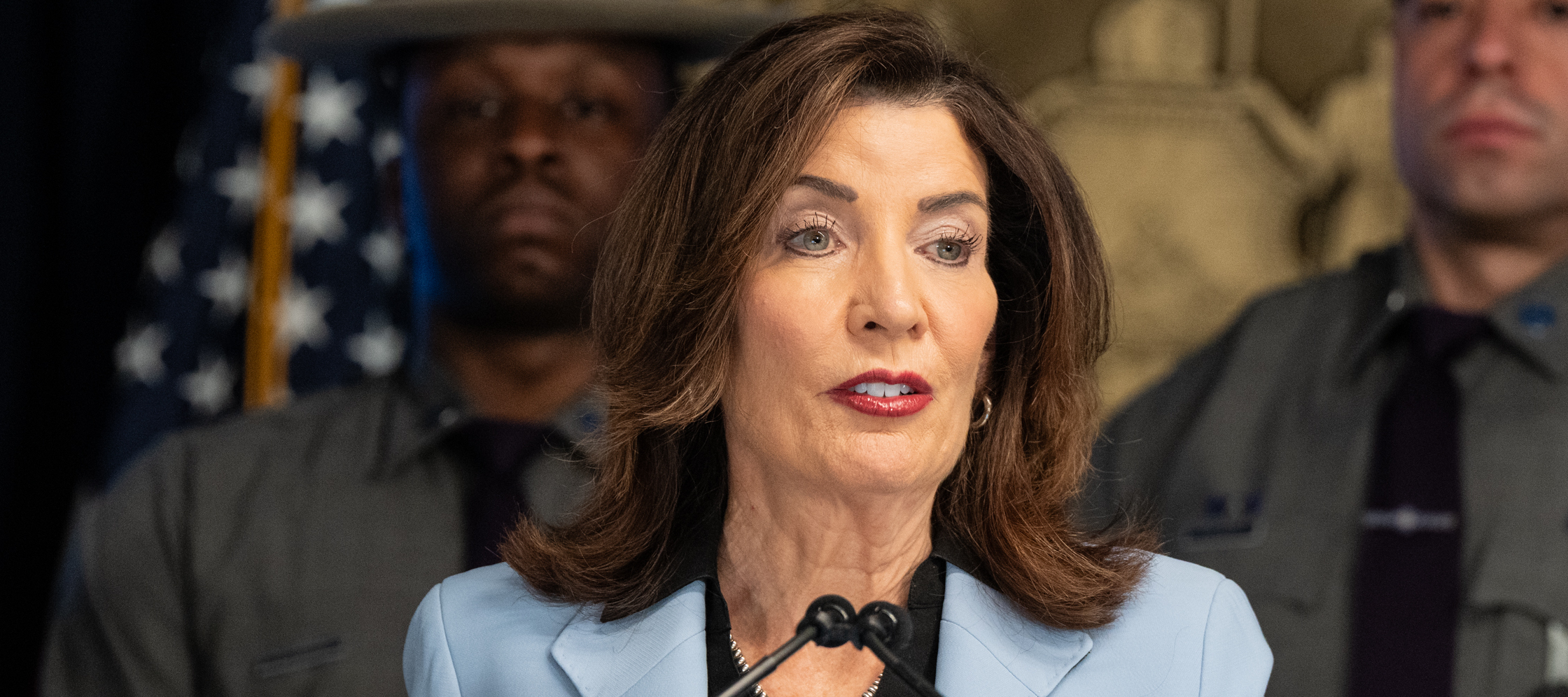 Kathy Hochul speaks at a podium, flanked by two NY state troopers.