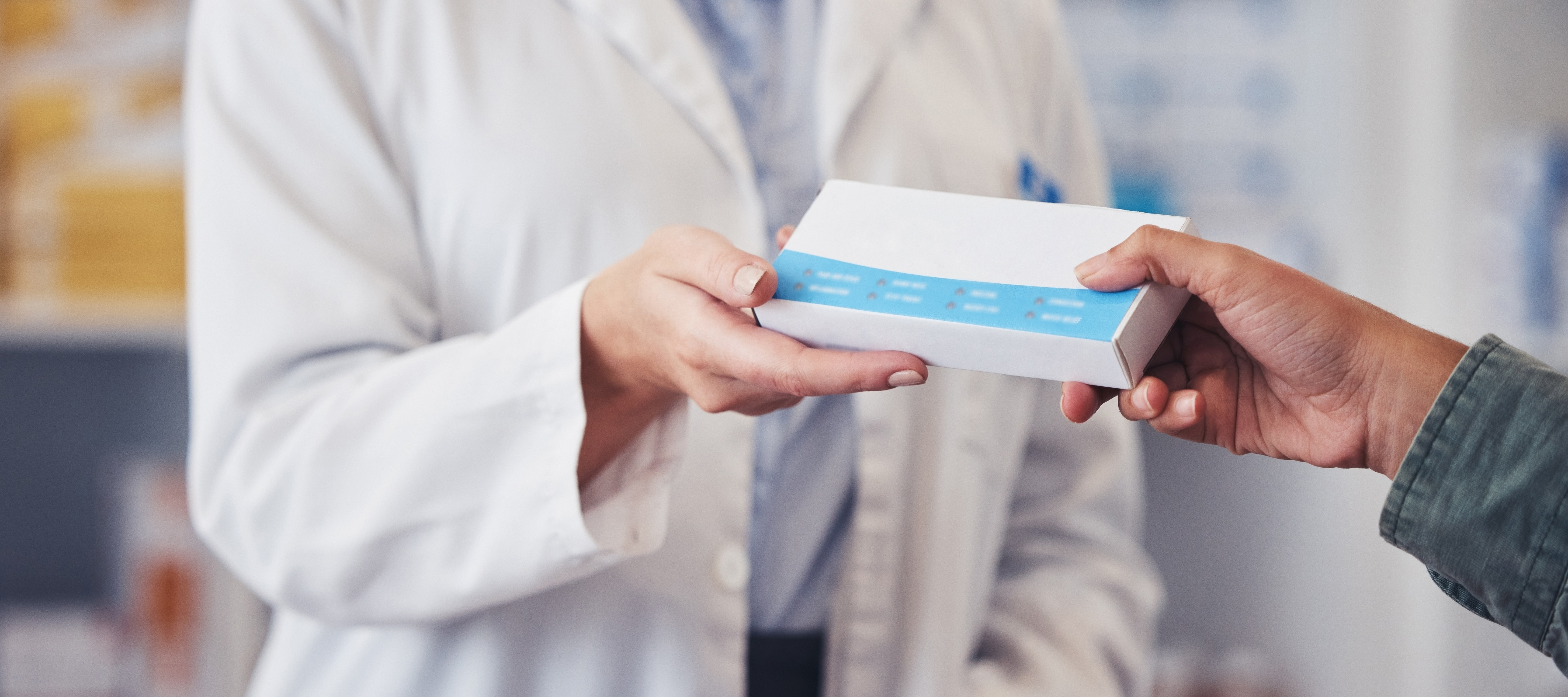 A pharmacist in a white lab coat hands a white and blue box to a customer over a wood counter in a pharmacy.