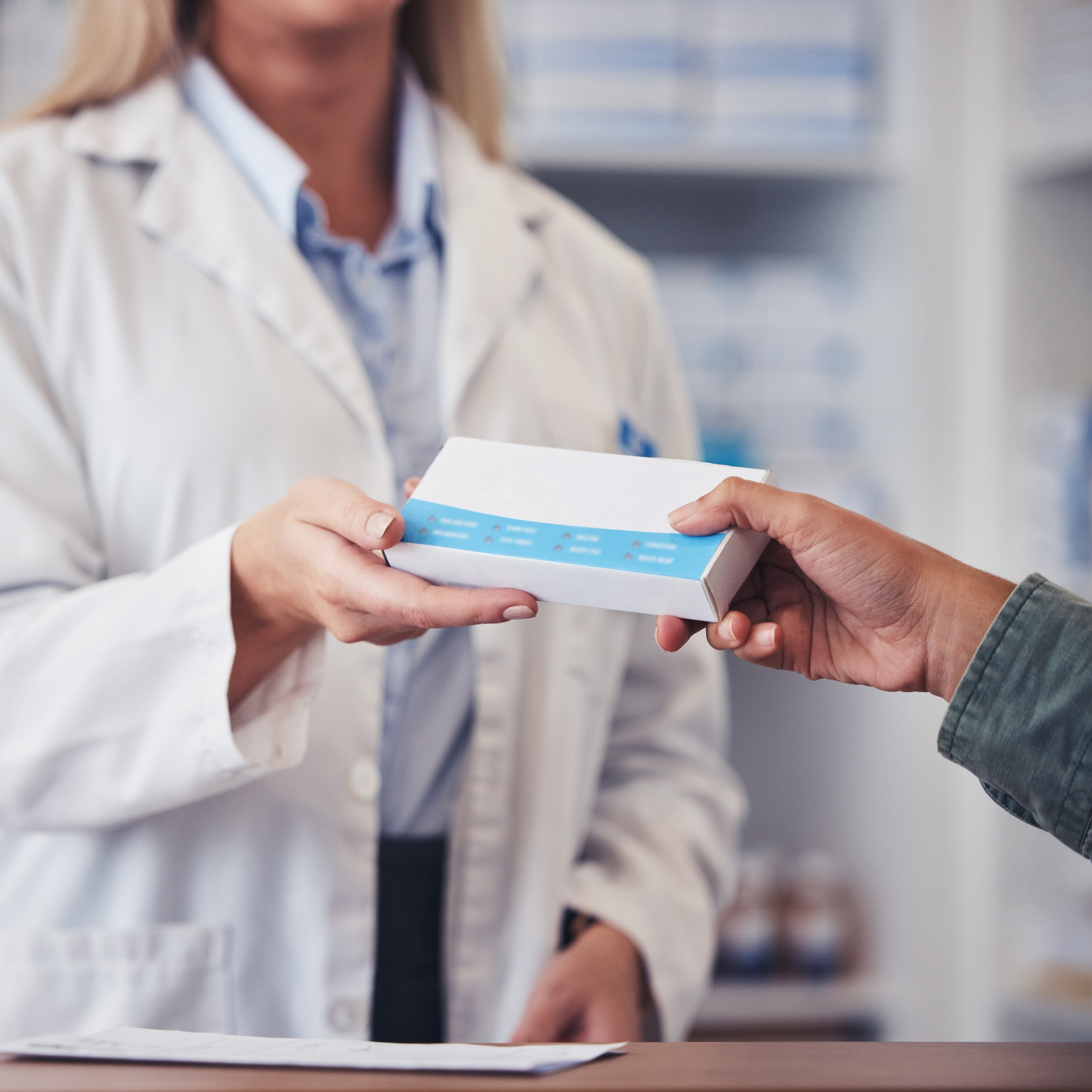 A pharmacist in a white lab coat hands a white and blue box to a customer over a wood counter in a pharmacy.