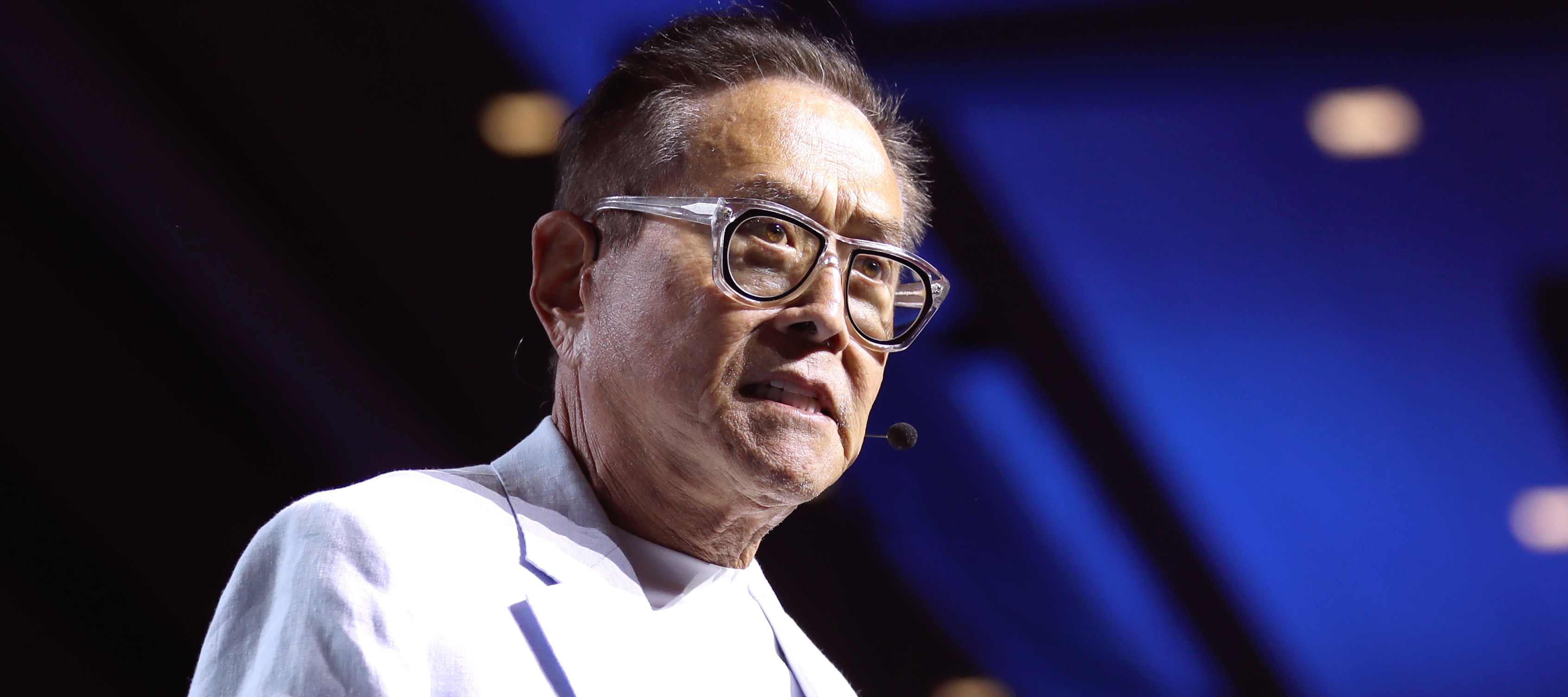 Robert Kiyosaki stands on a dark stage in an immaculate white suit.