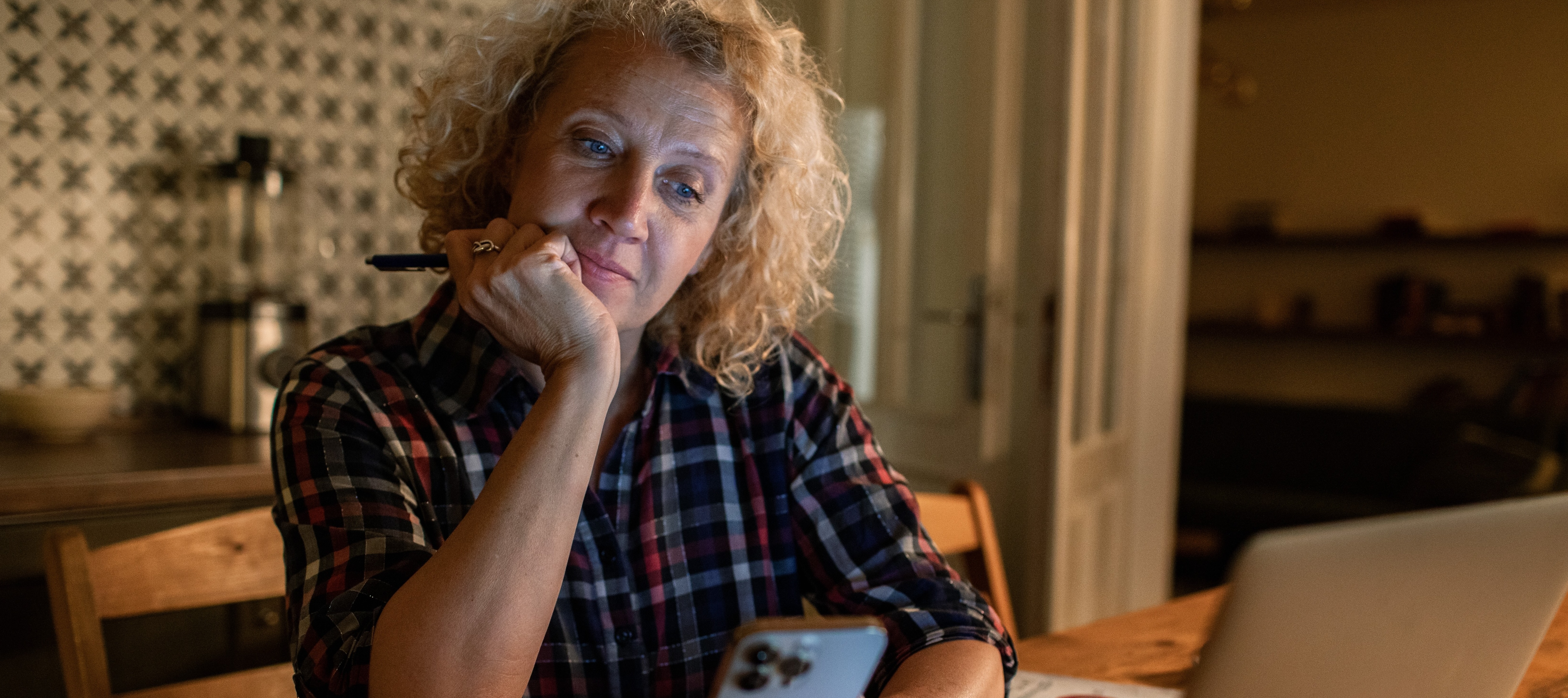 A blonde woman in a flannel work shirt sits in the dim light of her kitchen thinking deeply.