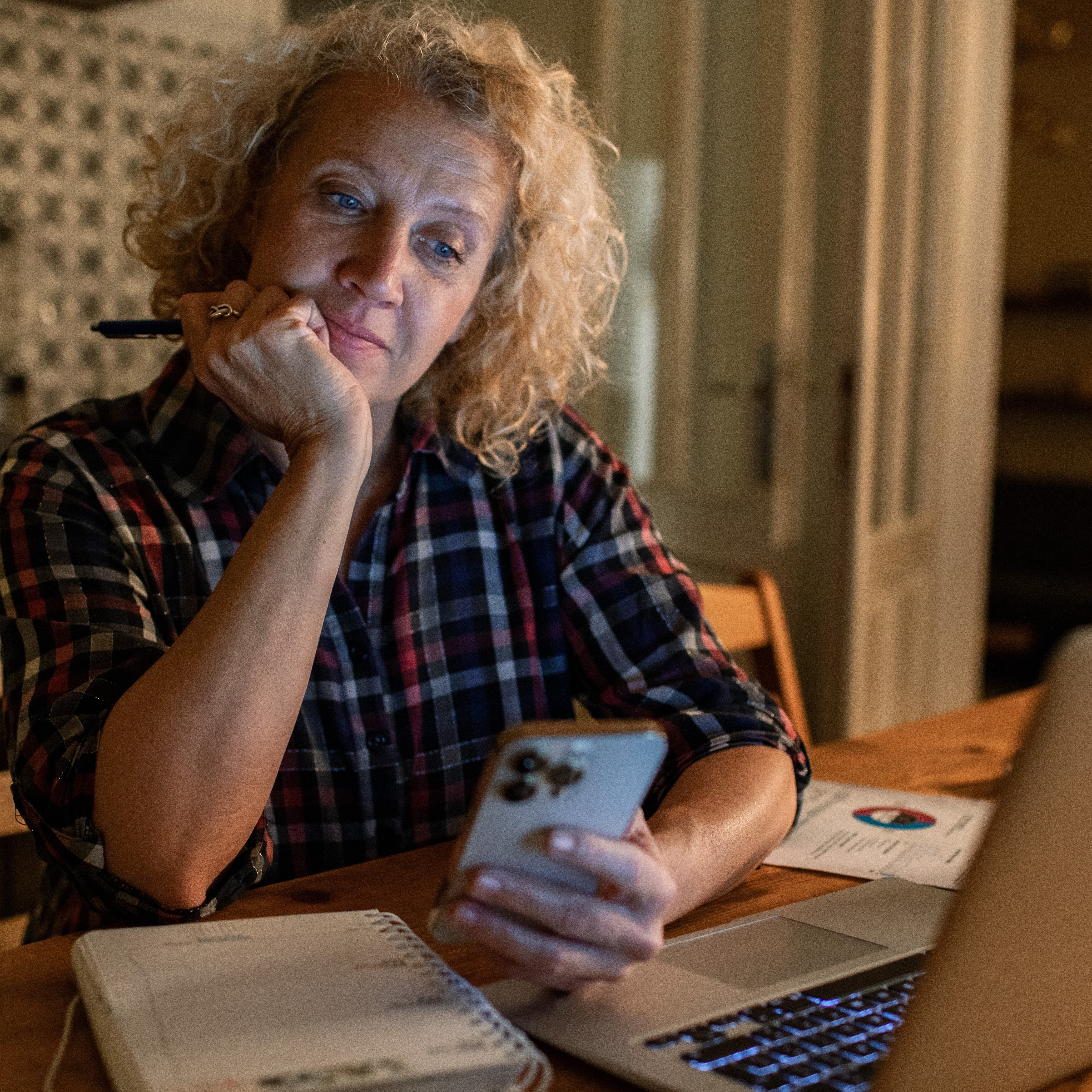 A blonde woman in a flannel work shirt sits in the dim light of her kitchen thinking deeply.