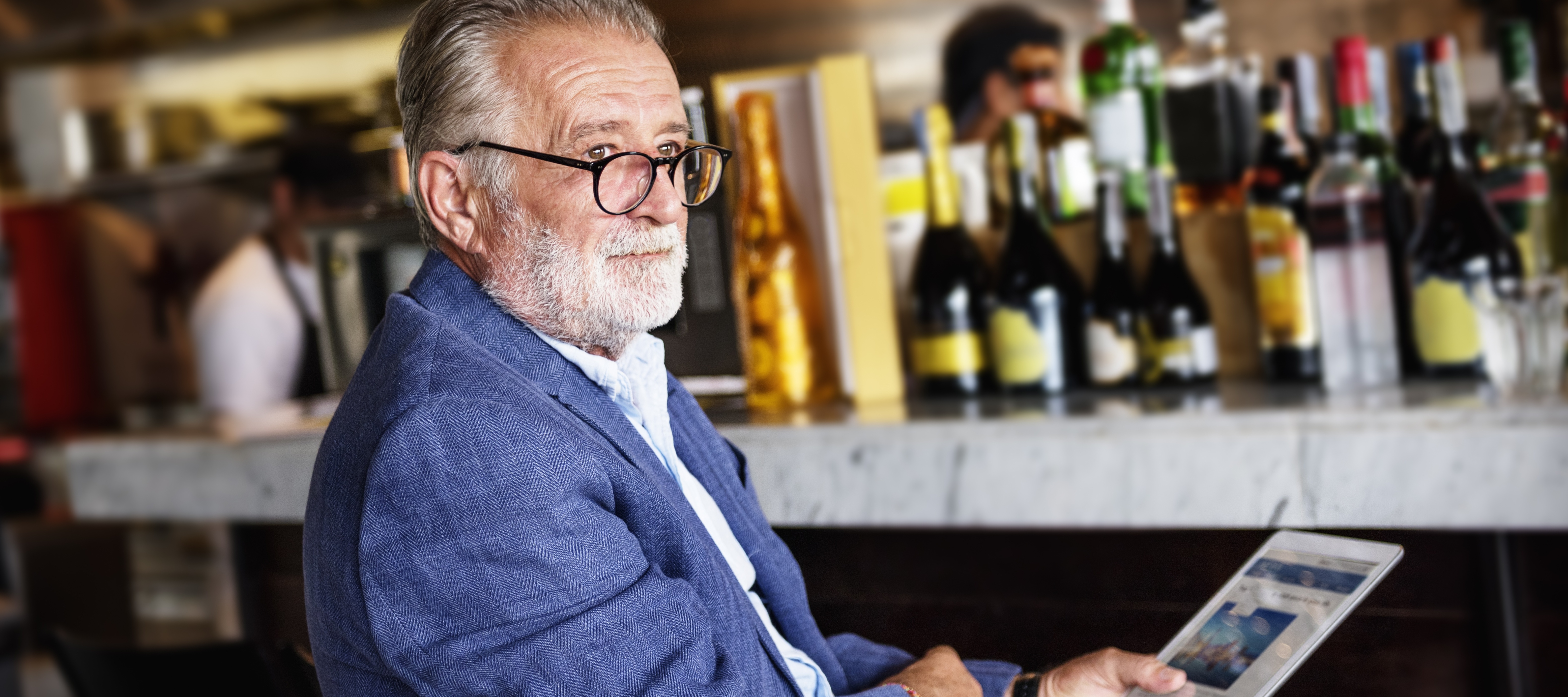 An older man sits at a bar, looking over a pair of thick rimmed glasses and into the middle distance.