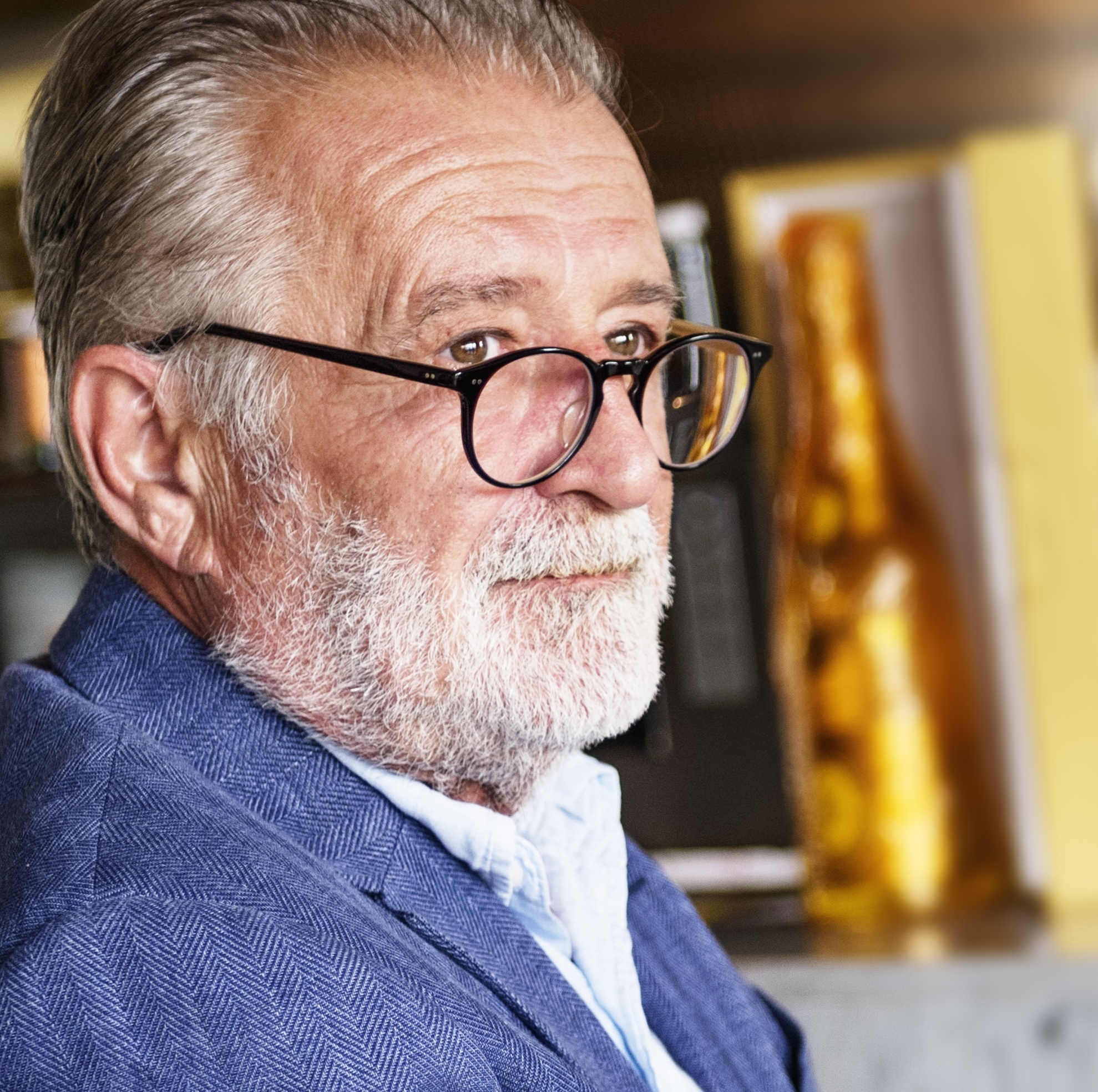 An older man sits at a bar, looking over a pair of thick rimmed glasses and into the middle distance.