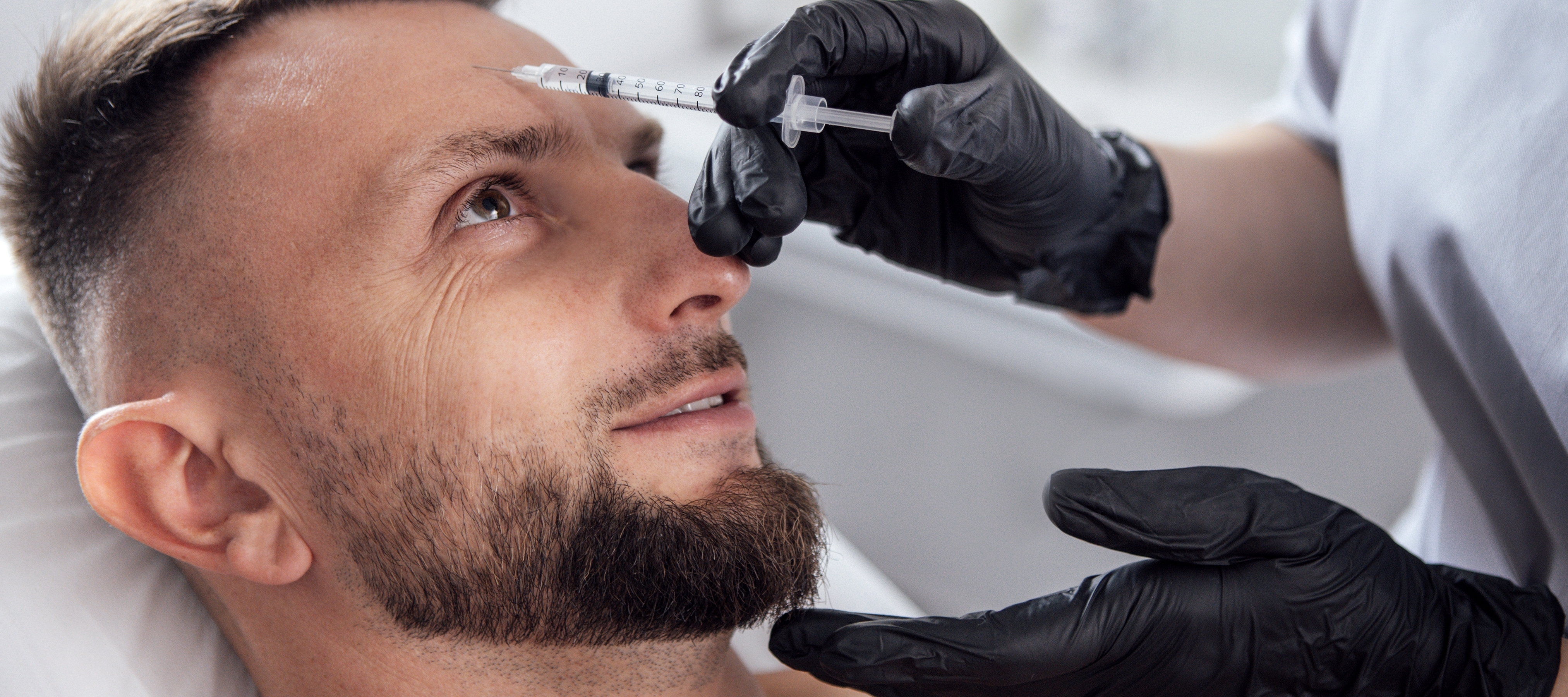 Male patient receiving cosmetic treatment with syringe by a healthcare professional in a clinical setting, wearing gloves.