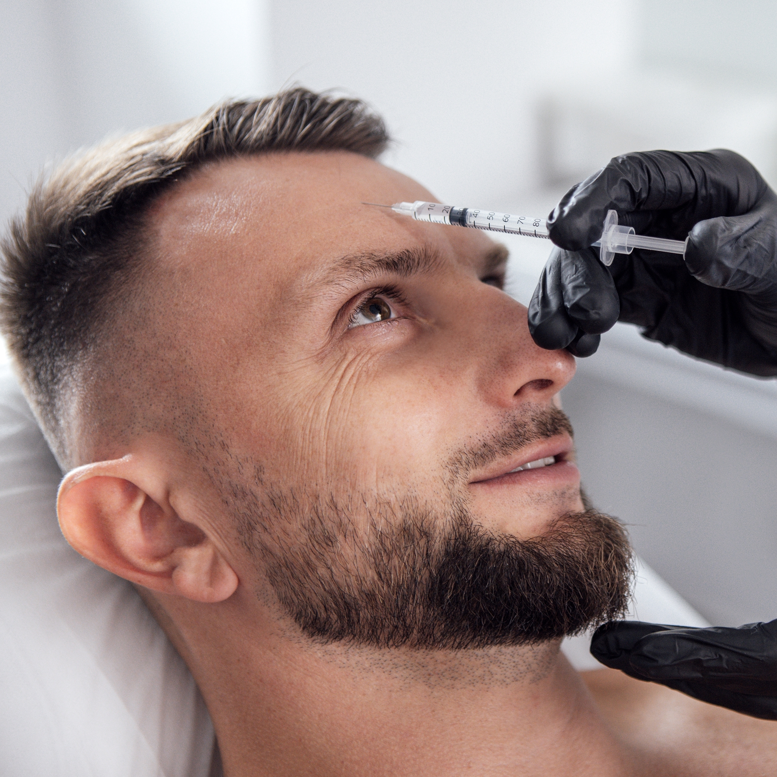 Male patient receiving cosmetic treatment with syringe by a healthcare professional in a clinical setting, wearing gloves.