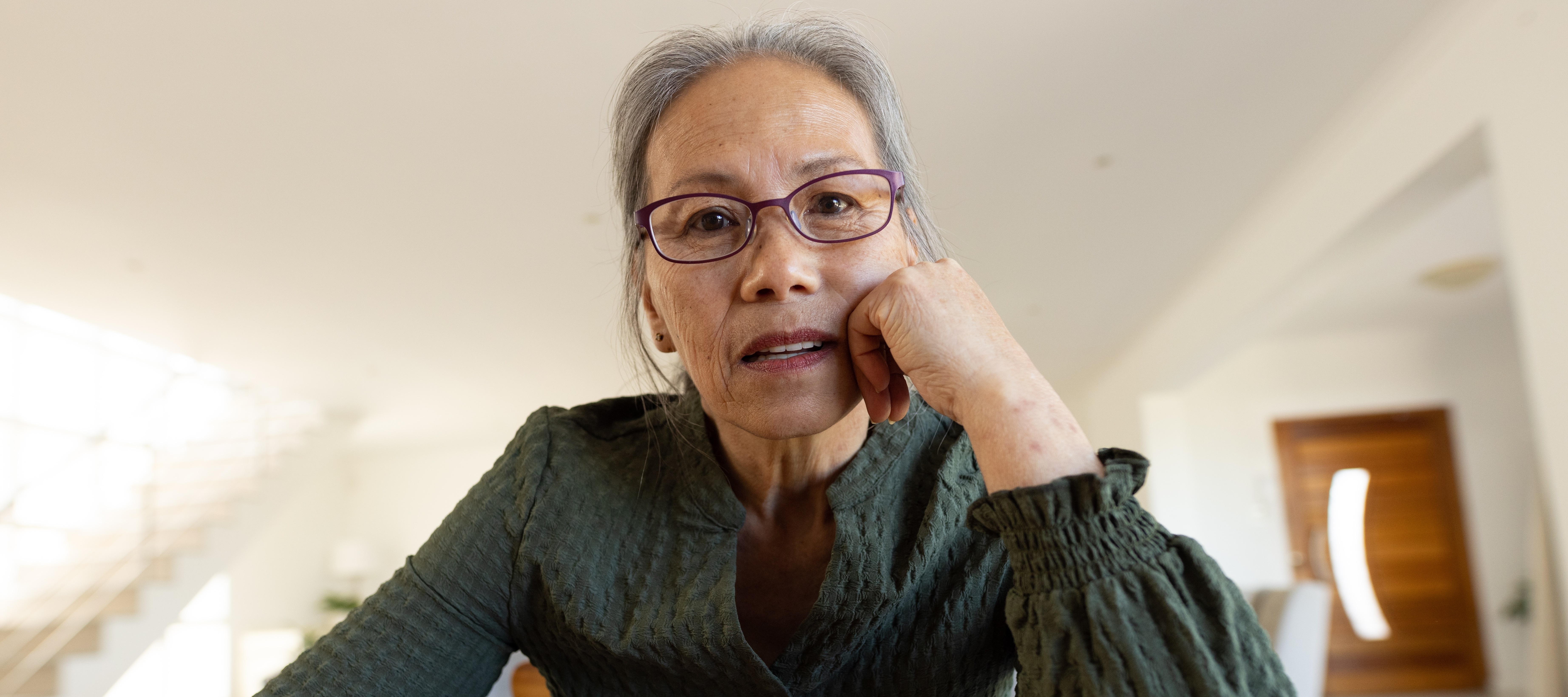 An older woman with short gray hair sits at a wooden table, resting her face on her hand.