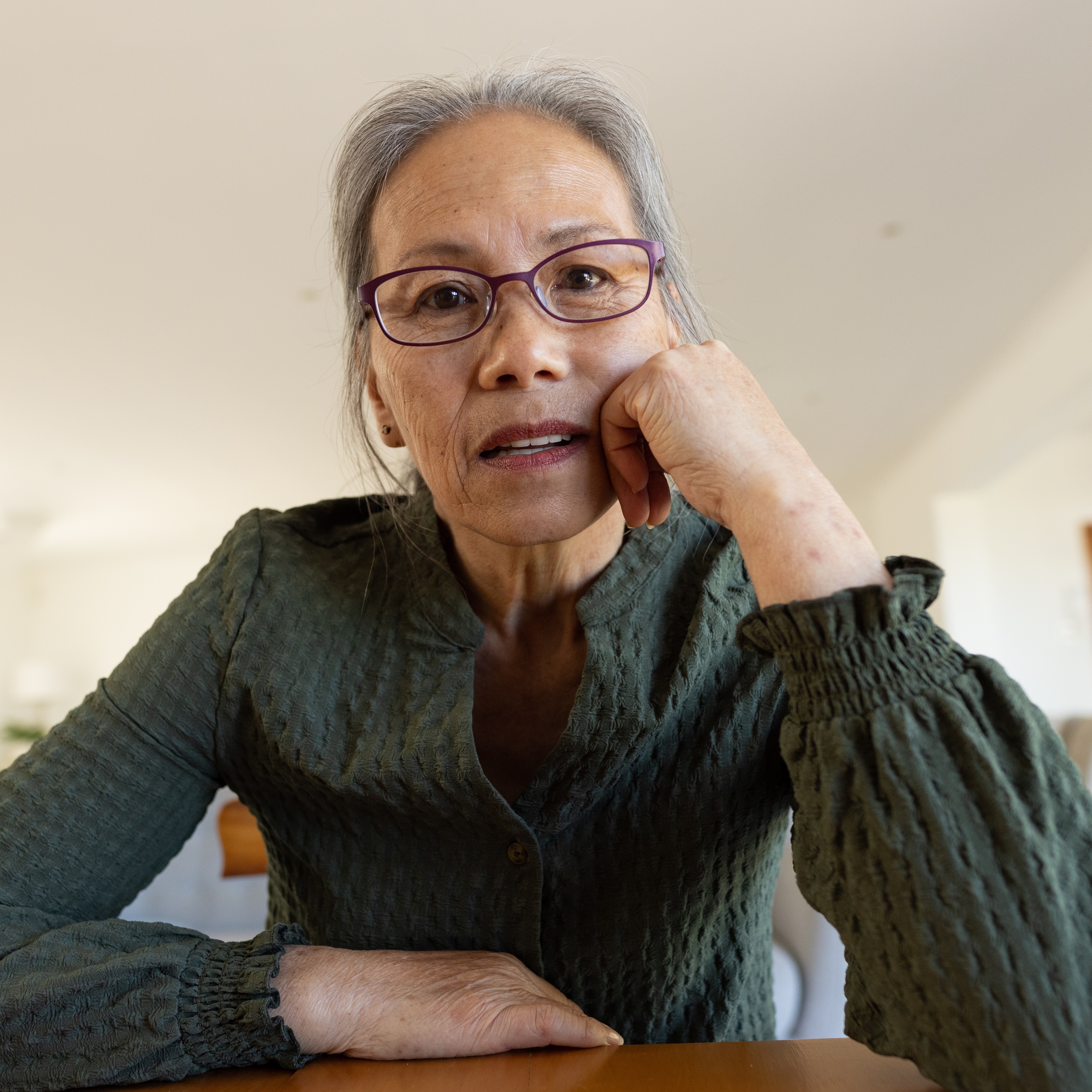An older woman with short gray hair sits at a wooden table, resting her face on her hand.