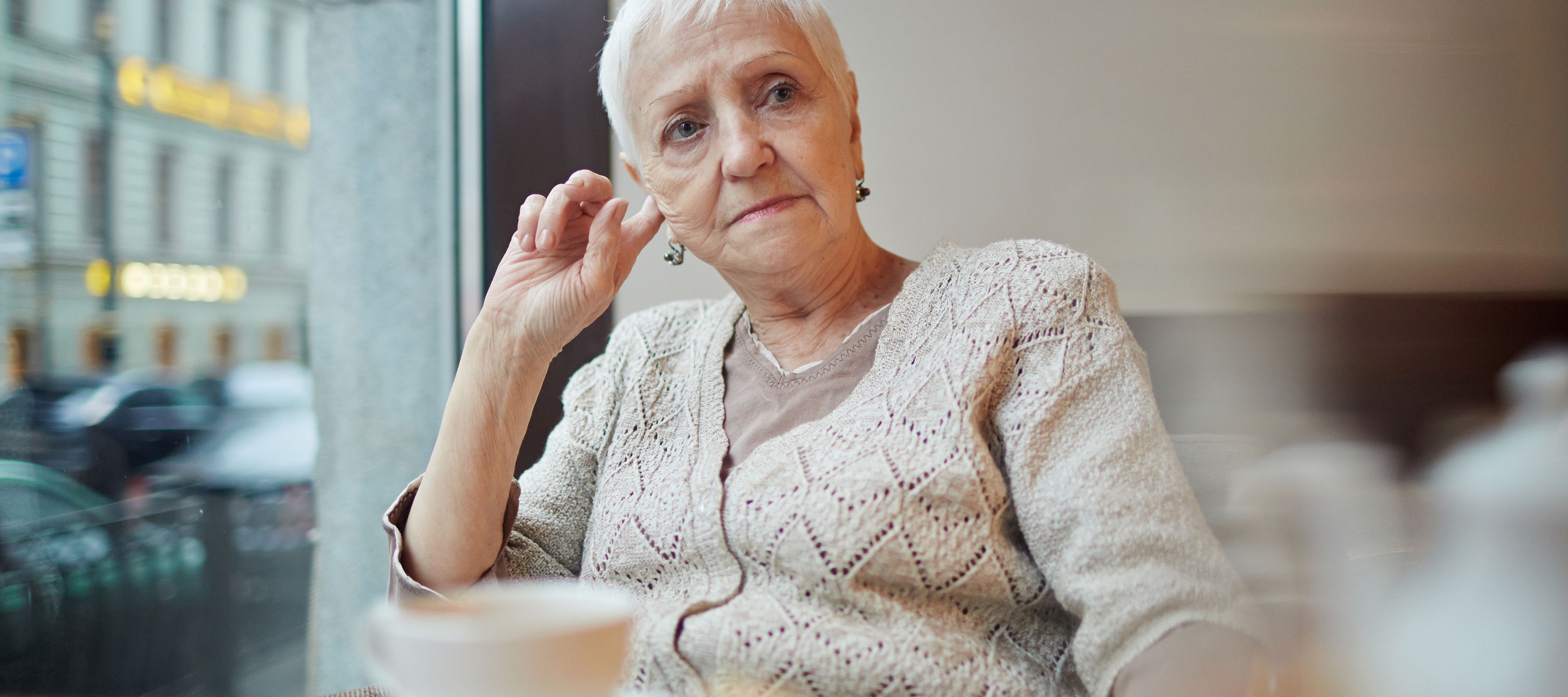 An older woman sits in a cafe with her coffee and a pensive look on her face.