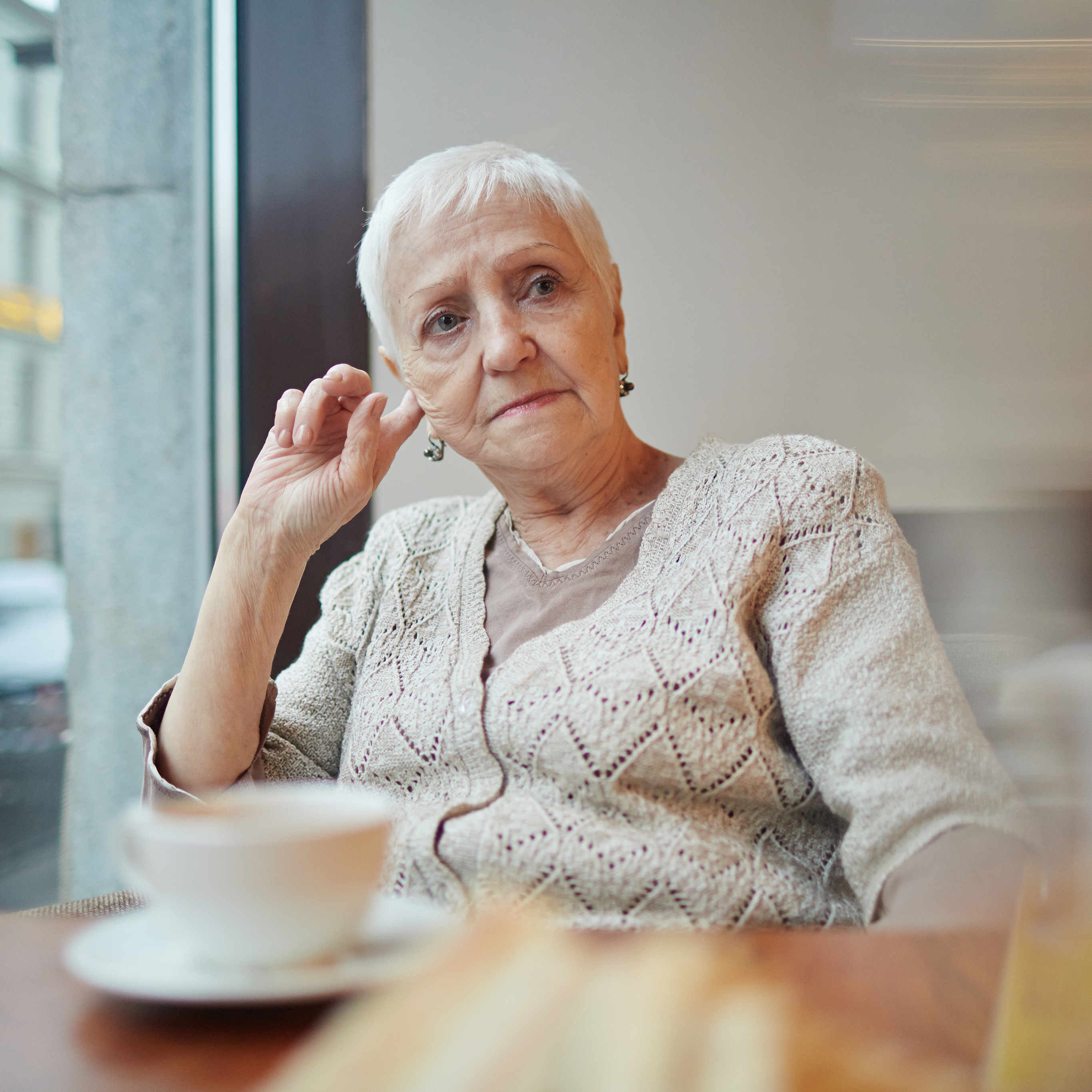 An older woman sits in a cafe with her coffee and a pensive look on her face.