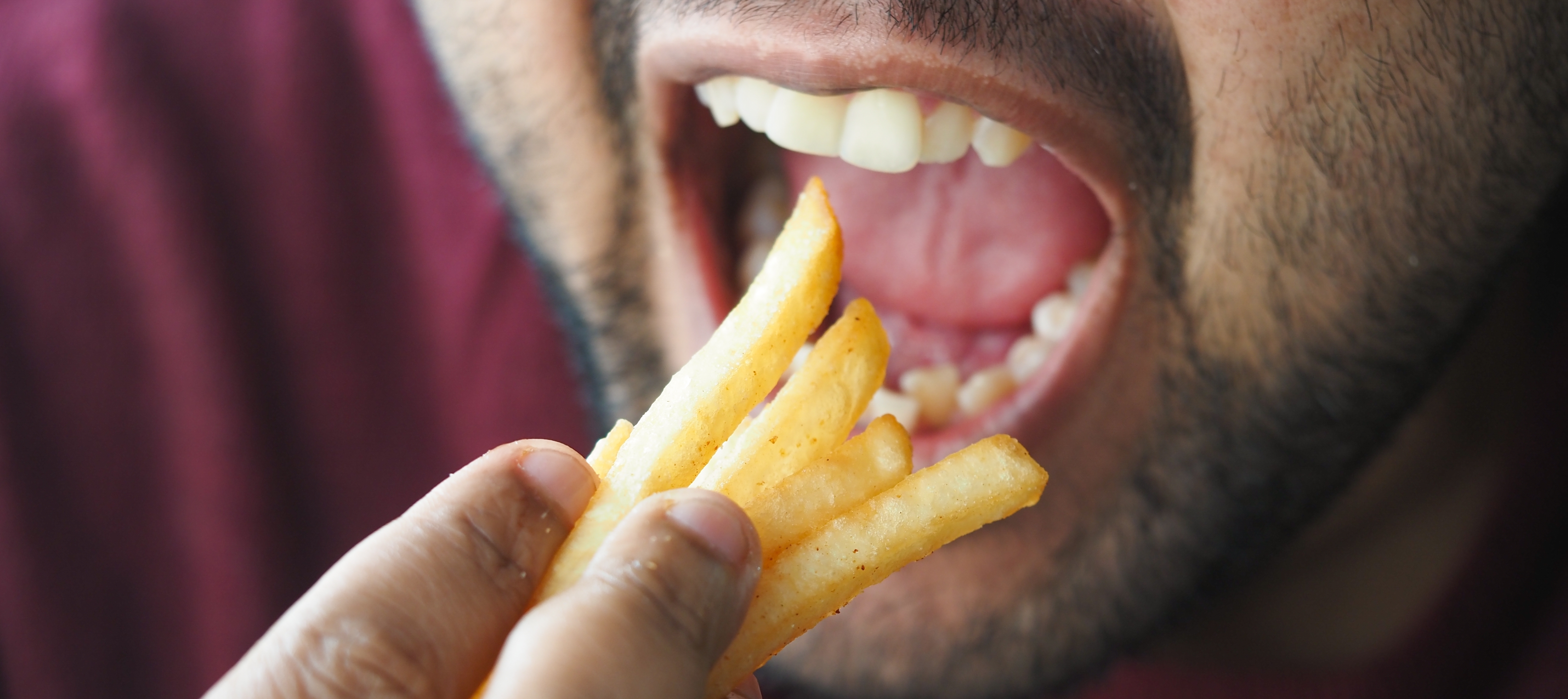 A close up of a man's mouth as he eats a handful of french fries.