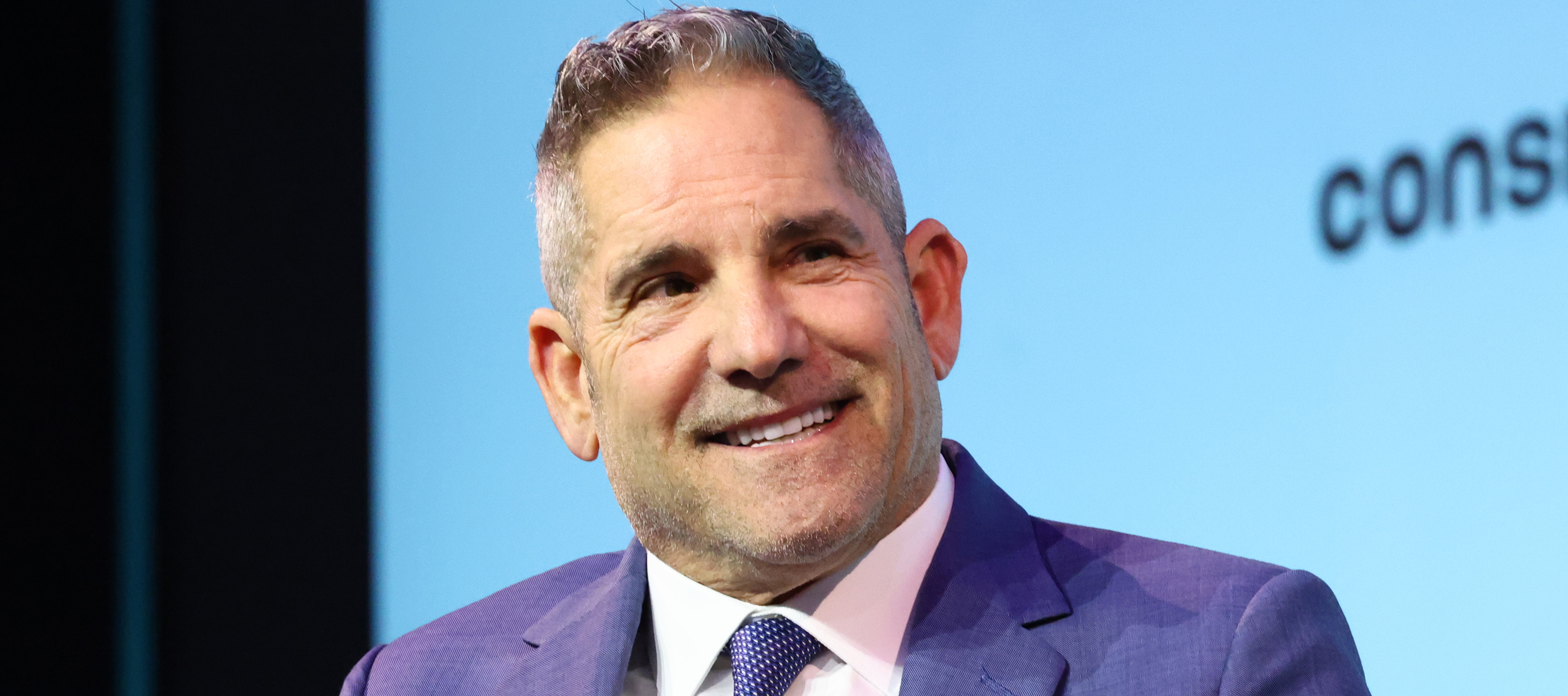 Grant Cardone smiles while speaking on stage while wearing a blue jacket and tie.
