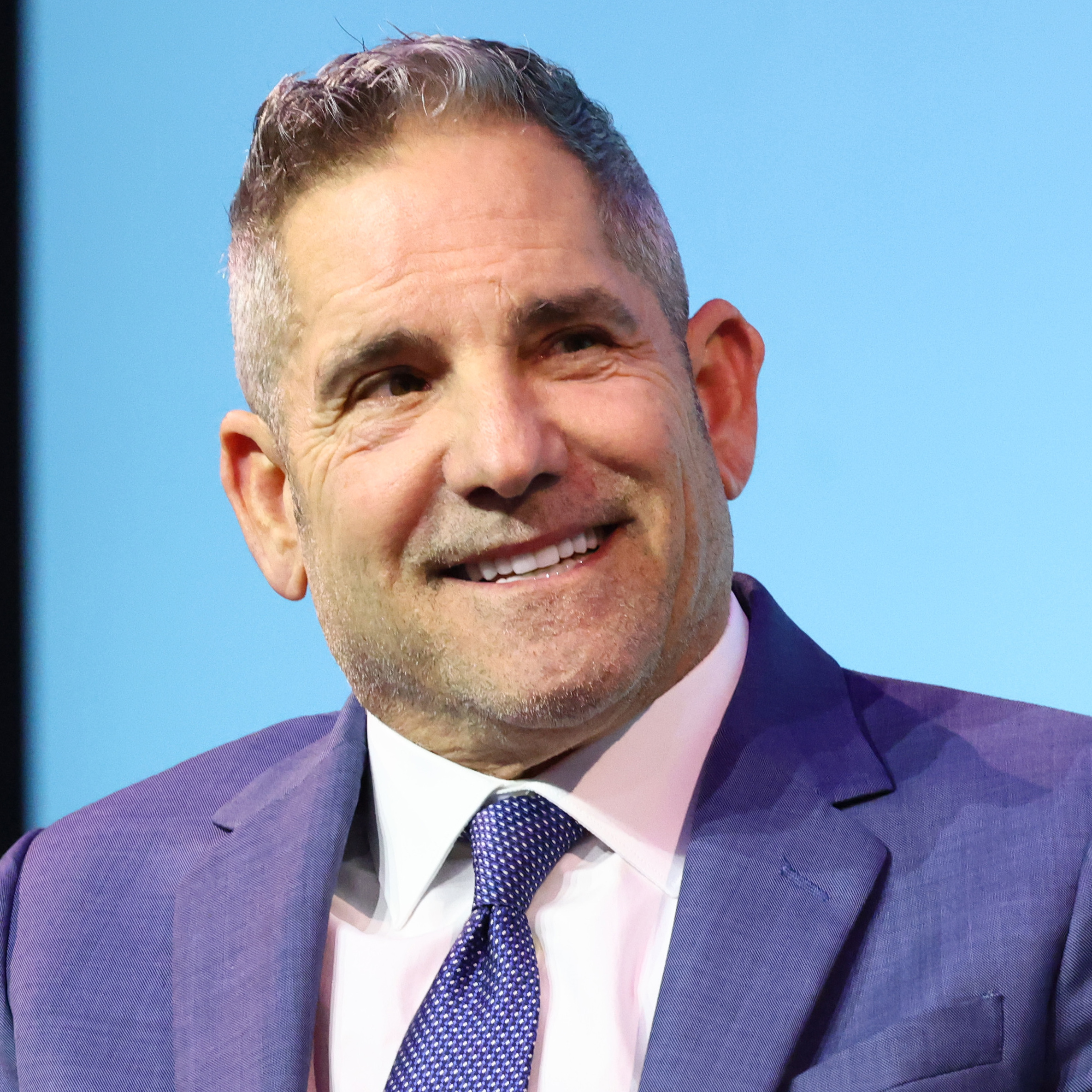Grant Cardone smiles while speaking on stage while wearing a blue jacket and tie.