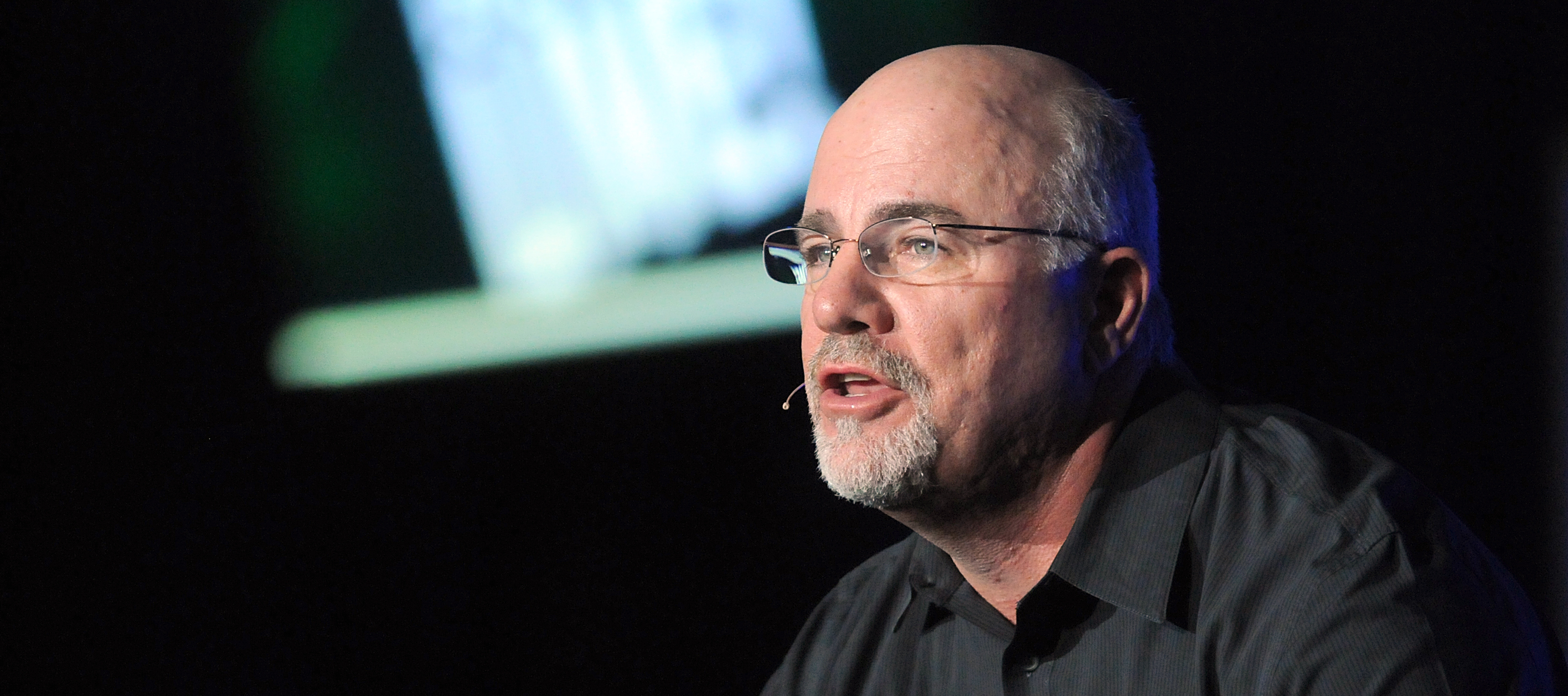 Dave Ramsey spreads his hands dramatically on stage in a black button up shirt.
