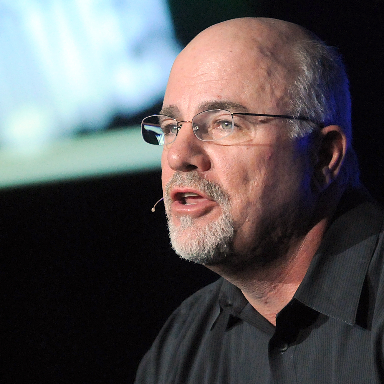 Dave Ramsey spreads his hands dramatically on stage in a black button up shirt.