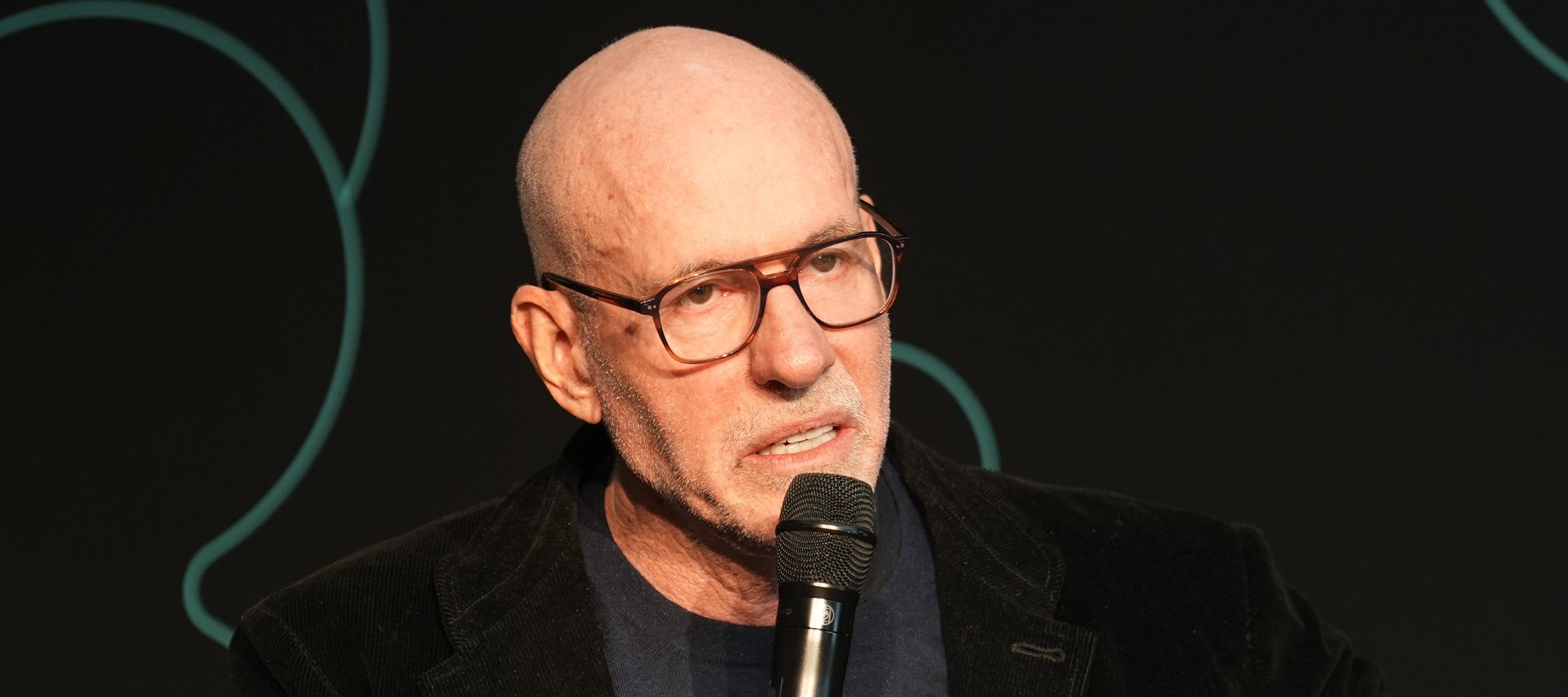 Scott Galloway, also known as Prof G, grimaces while speaking into a microphone and gesturing outward with one hand.