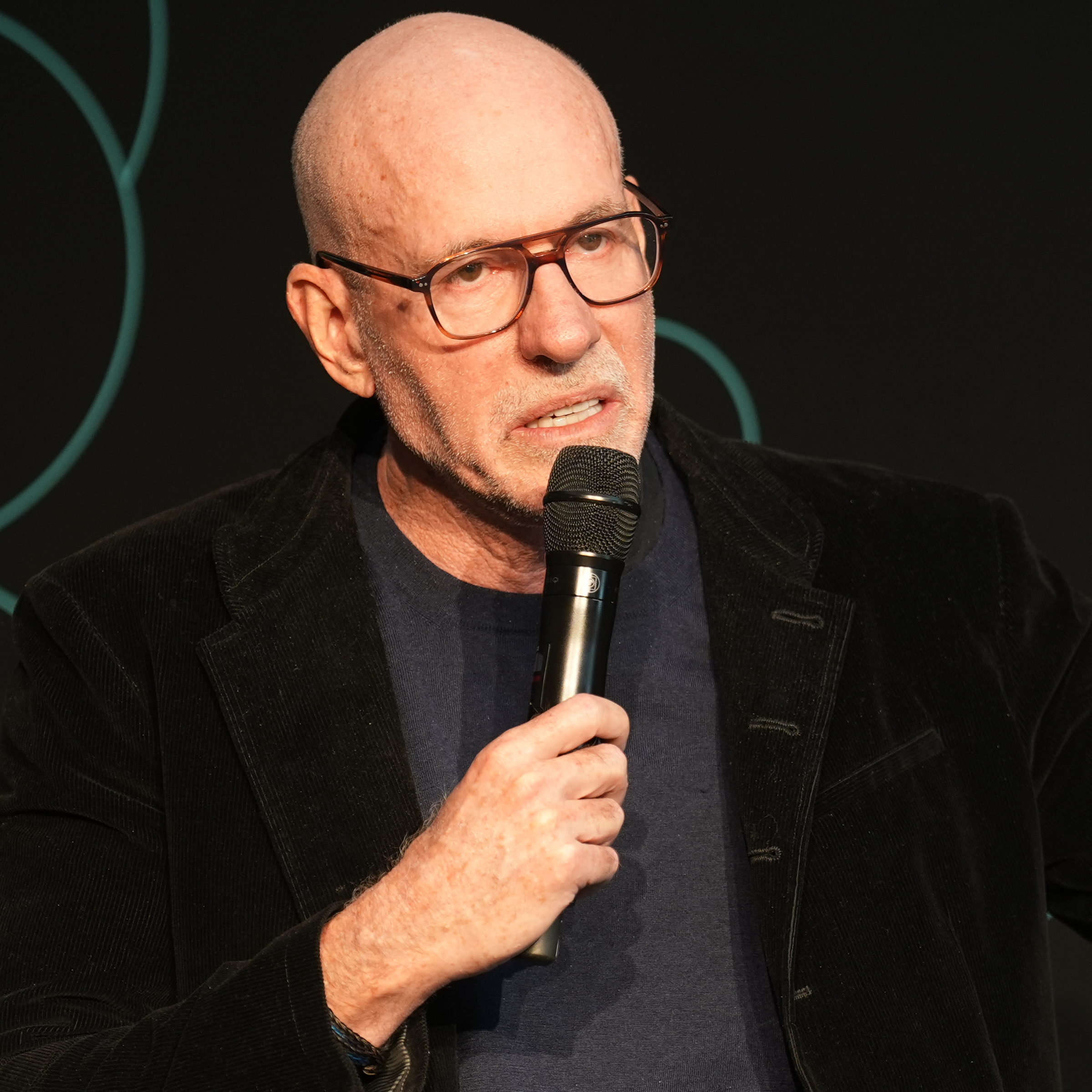 Scott Galloway, also known as Prof G, grimaces while speaking into a microphone and gesturing outward with one hand.