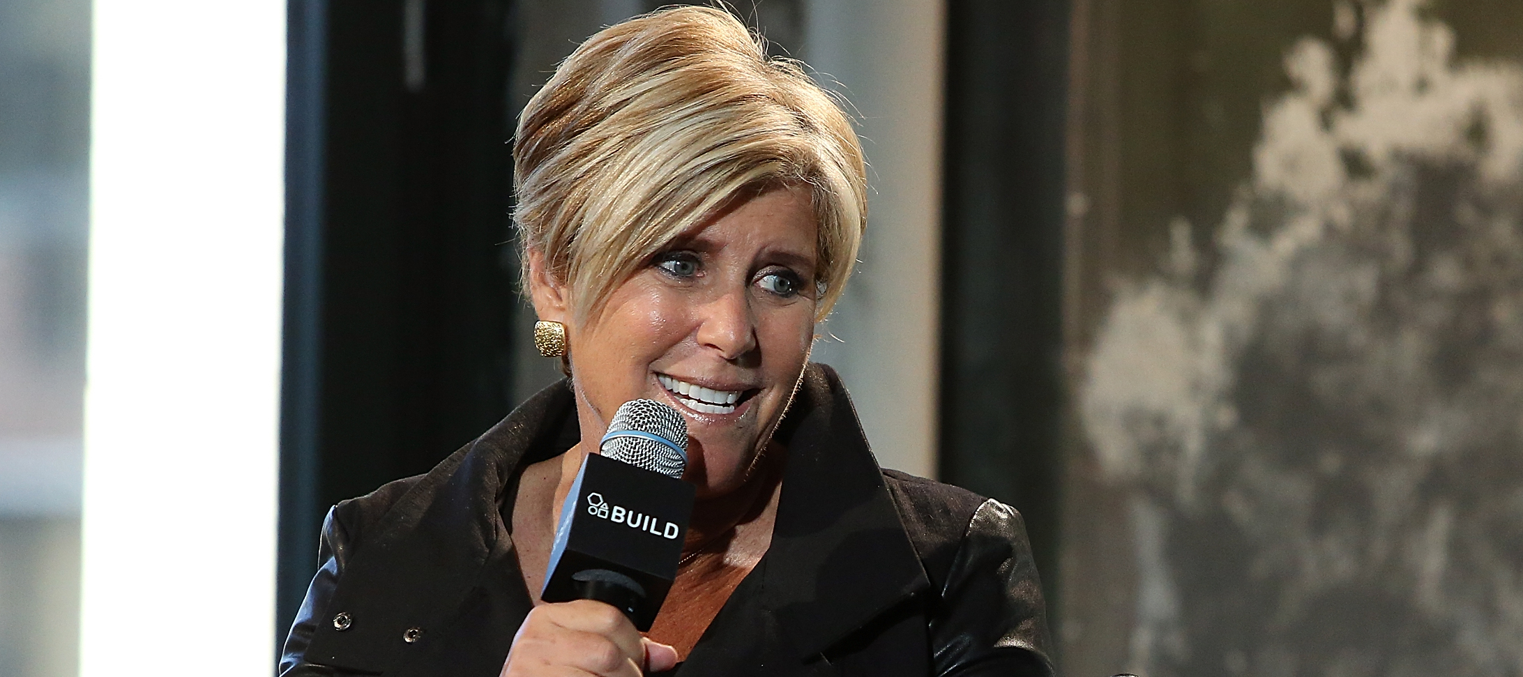 Suze Orman, wearing a black leather jacket, reclines in a chair and speaks into a microphone.