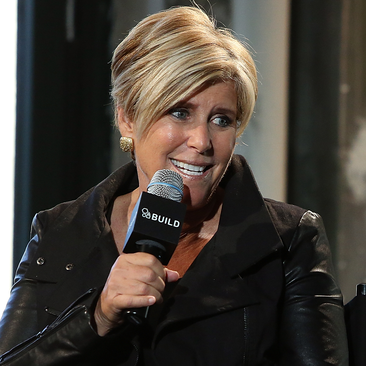 Suze Orman, wearing a black leather jacket, reclines in a chair and speaks into a microphone.