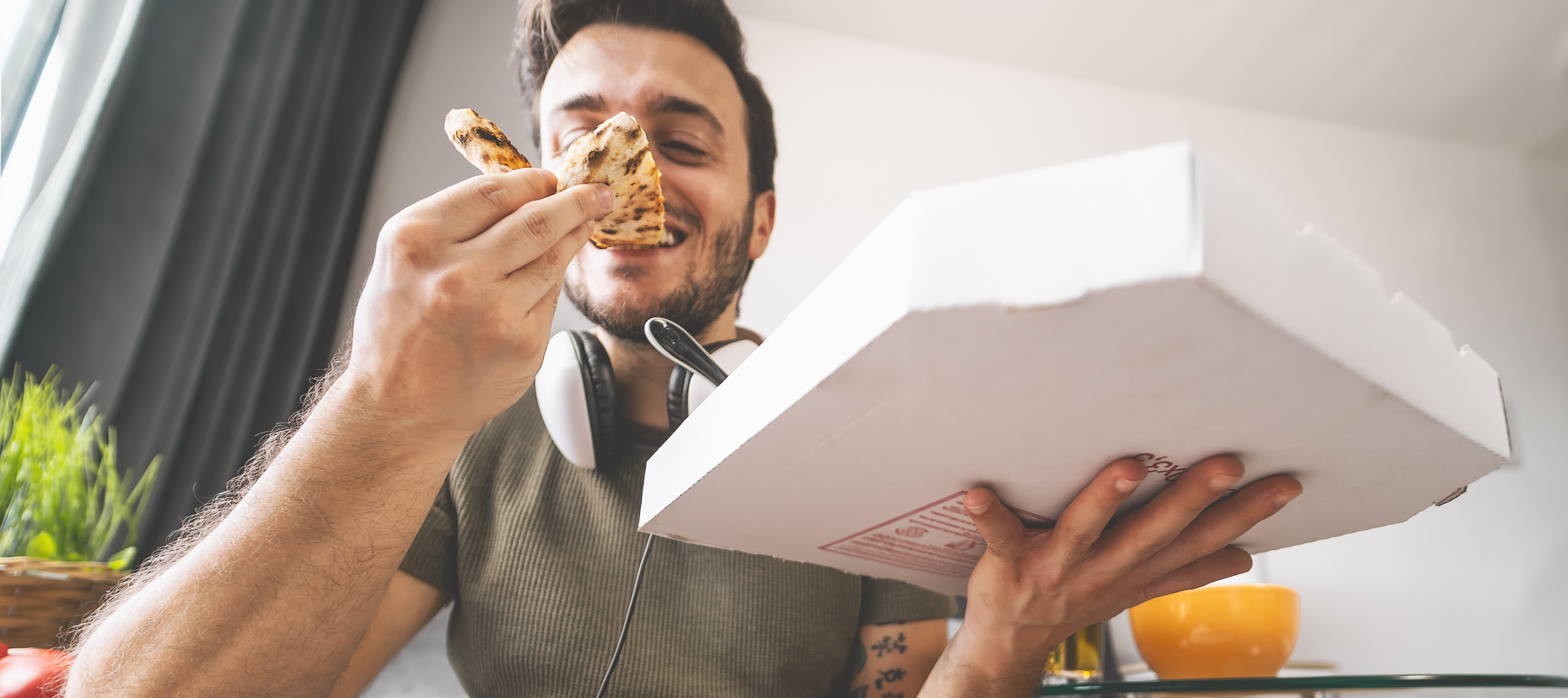 A smiling adult man with dark hair take a bit out of a slice of pizza in front of an open pizza box while sitting on the couch.