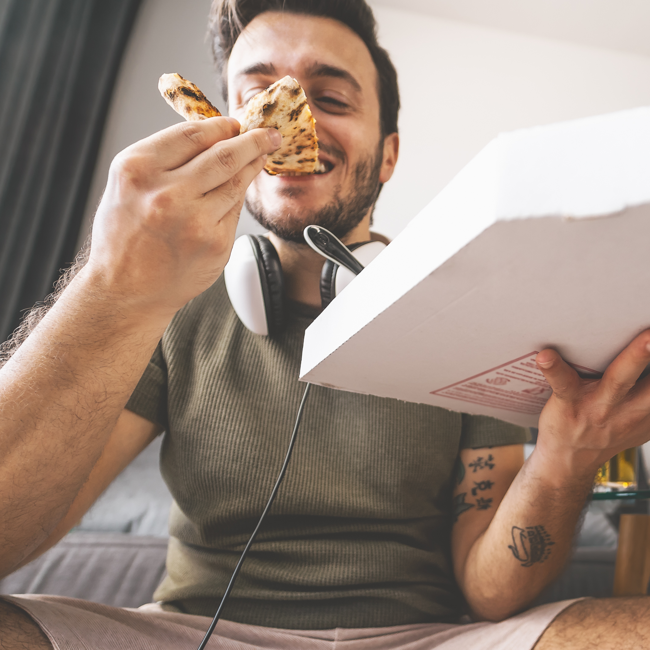 A smiling adult man with dark hair take a bit out of a slice of pizza in front of an open pizza box while sitting on the couch.