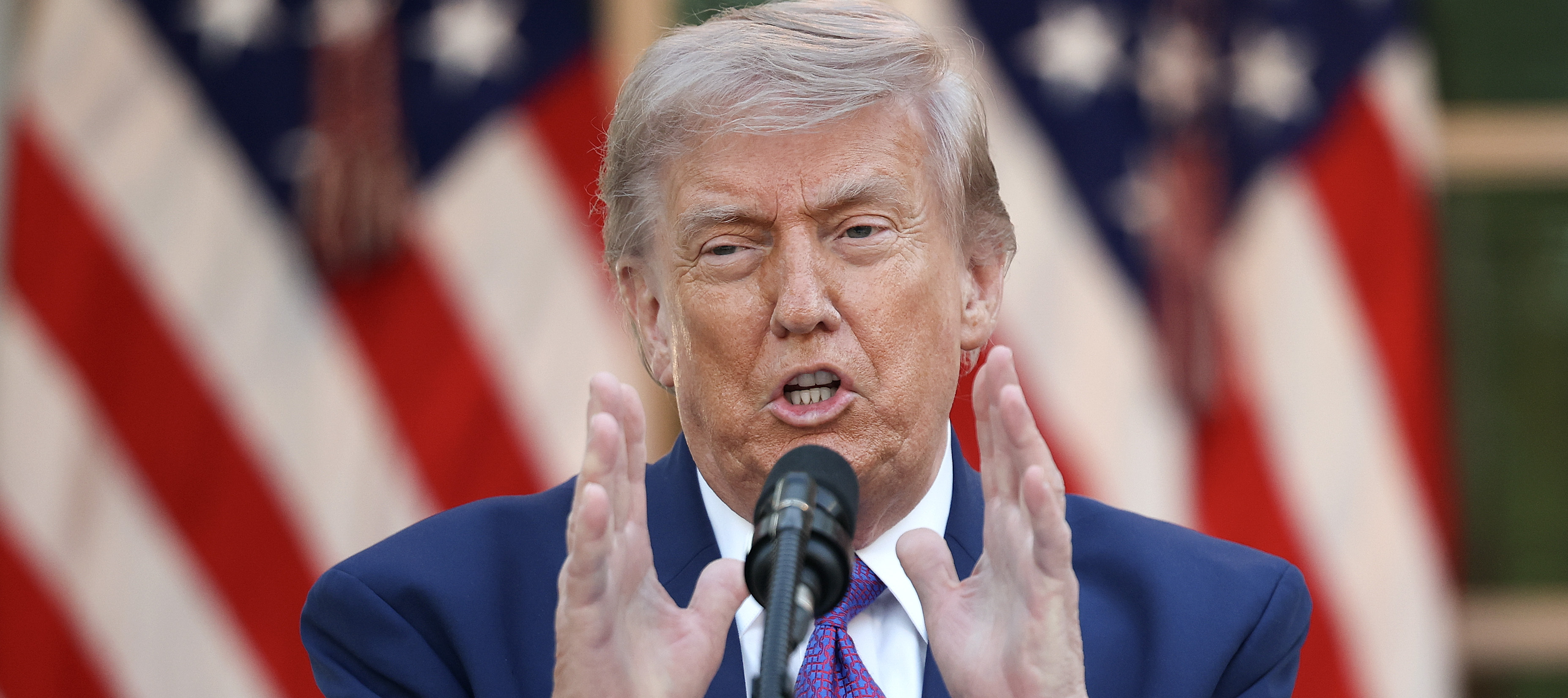 President Donald Trump speaks at a podium, holding up both hands.