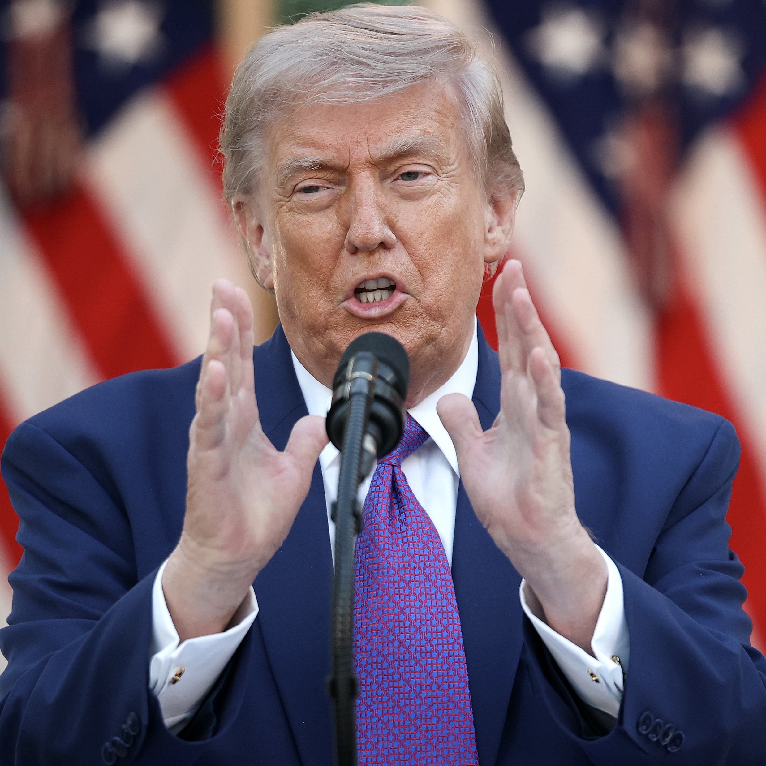 President Donald Trump speaks at a podium, holding up both hands.
