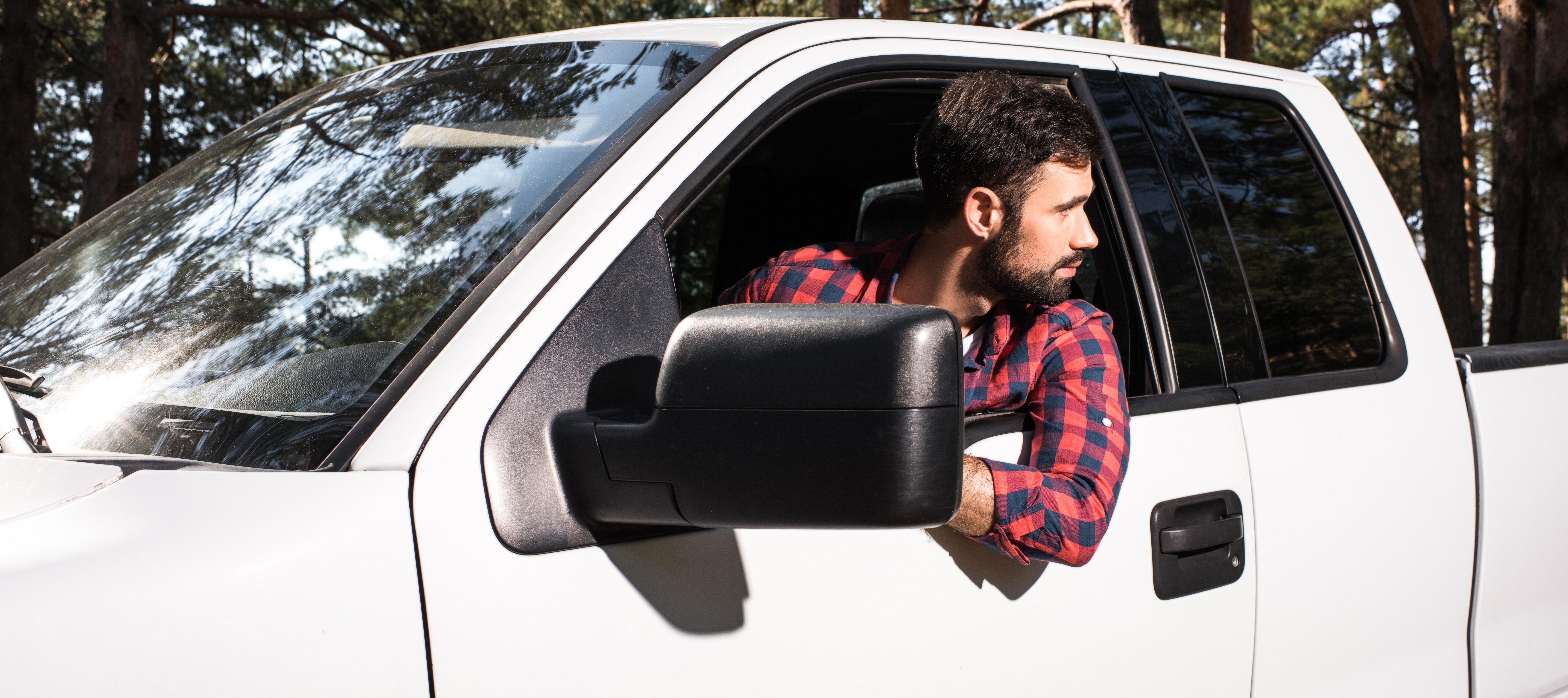 An adult man with short dark hair and a beard is seen inside the cab of a white pickup truck while backing it up, leaning out the window.