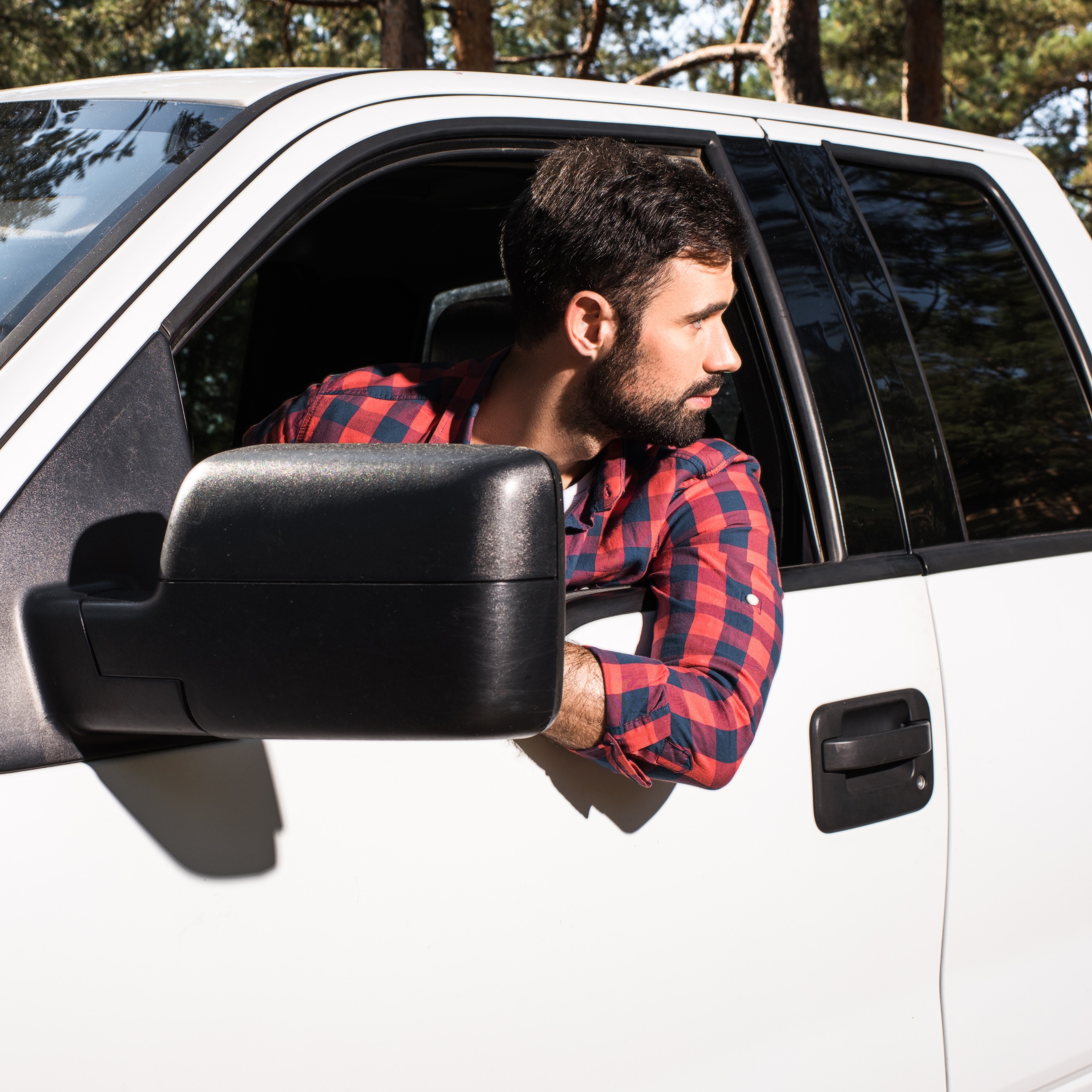 An adult man with short dark hair and a beard is seen inside the cab of a white pickup truck while backing it up, leaning out the window.