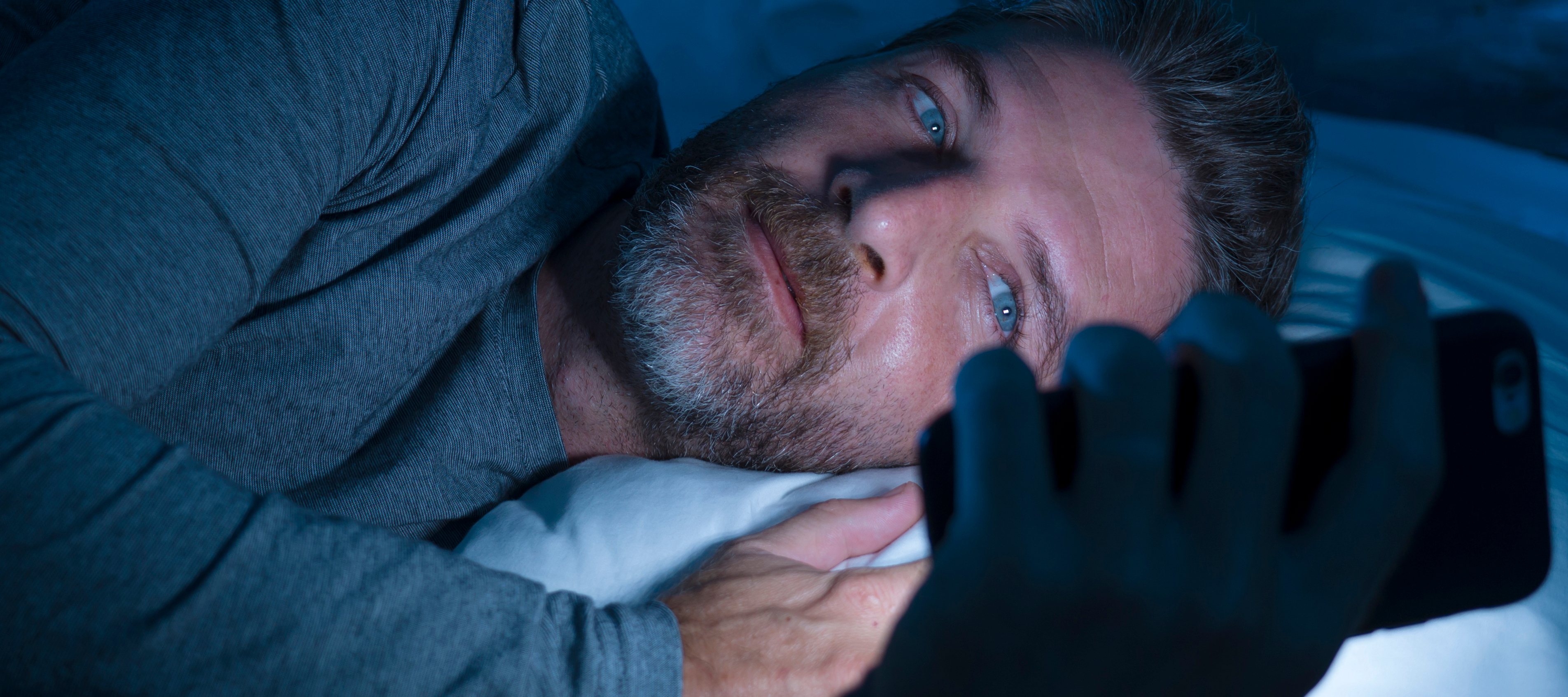 A man is scrolling his phone in bed because money worries are keeping him up at night.
