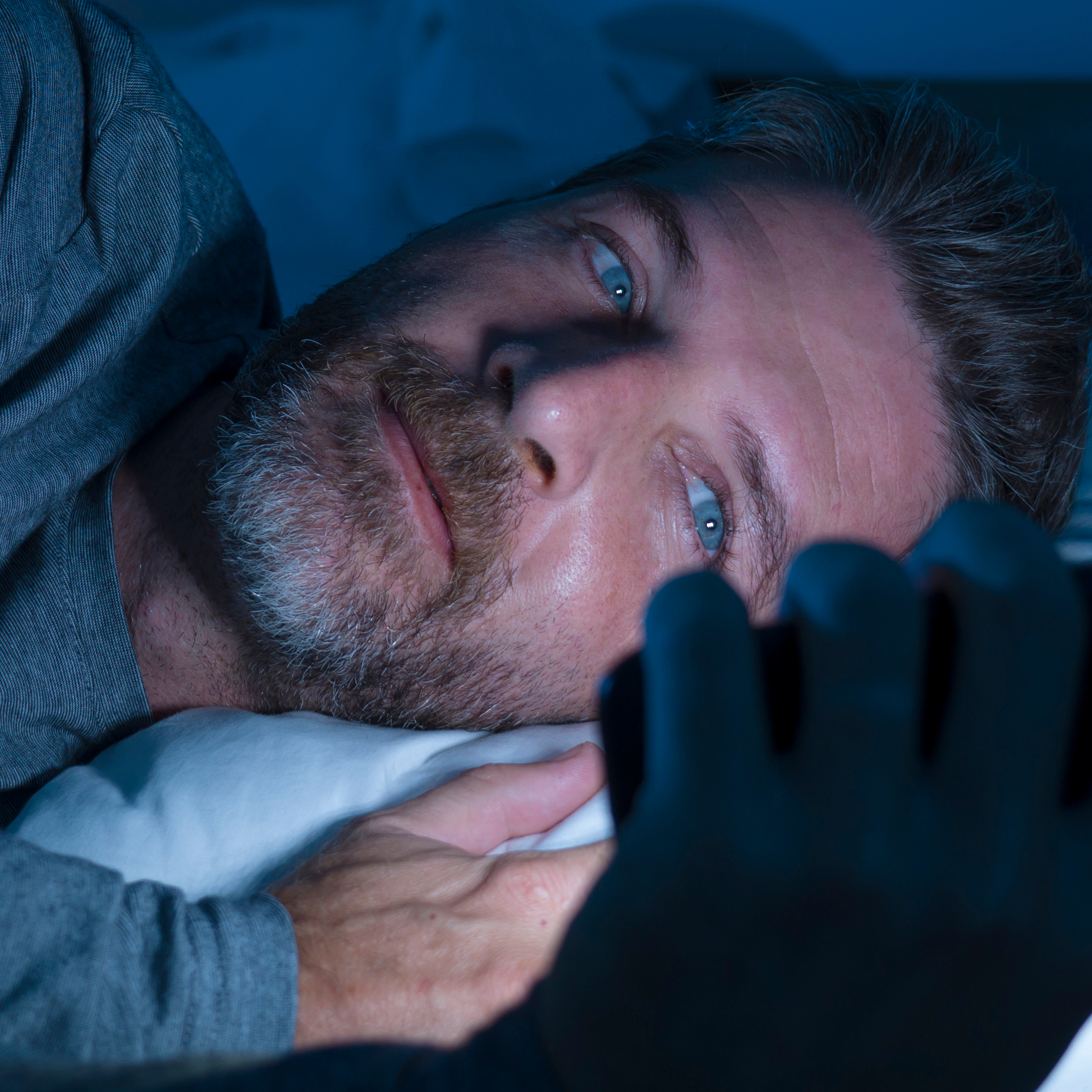 A man is scrolling his phone in bed because money worries are keeping him up at night.