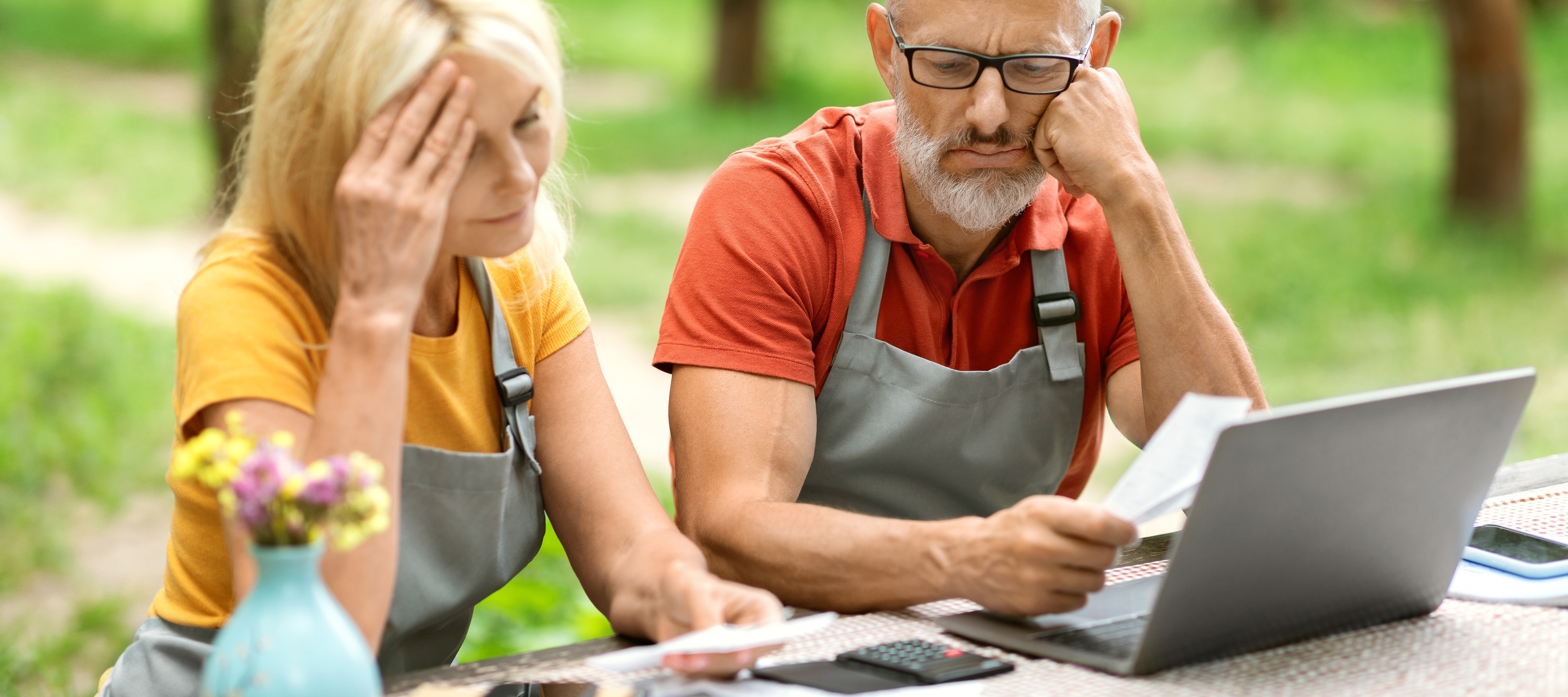 A blonde woman and a man with gray hair sit at a table outside, both wearing gray aprons, with concerned expressions.