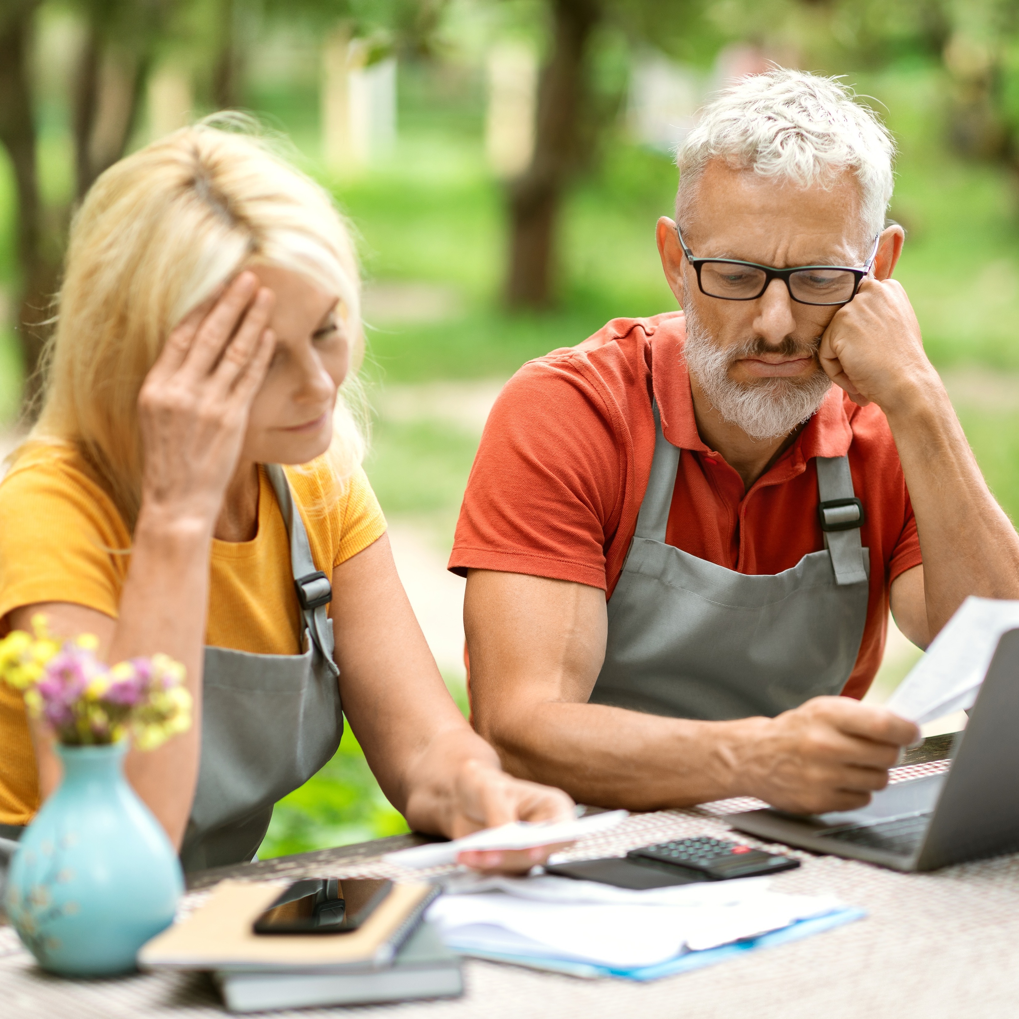 A blonde woman and a man with gray hair sit at a table outside, both wearing gray aprons, with concerned expressions.